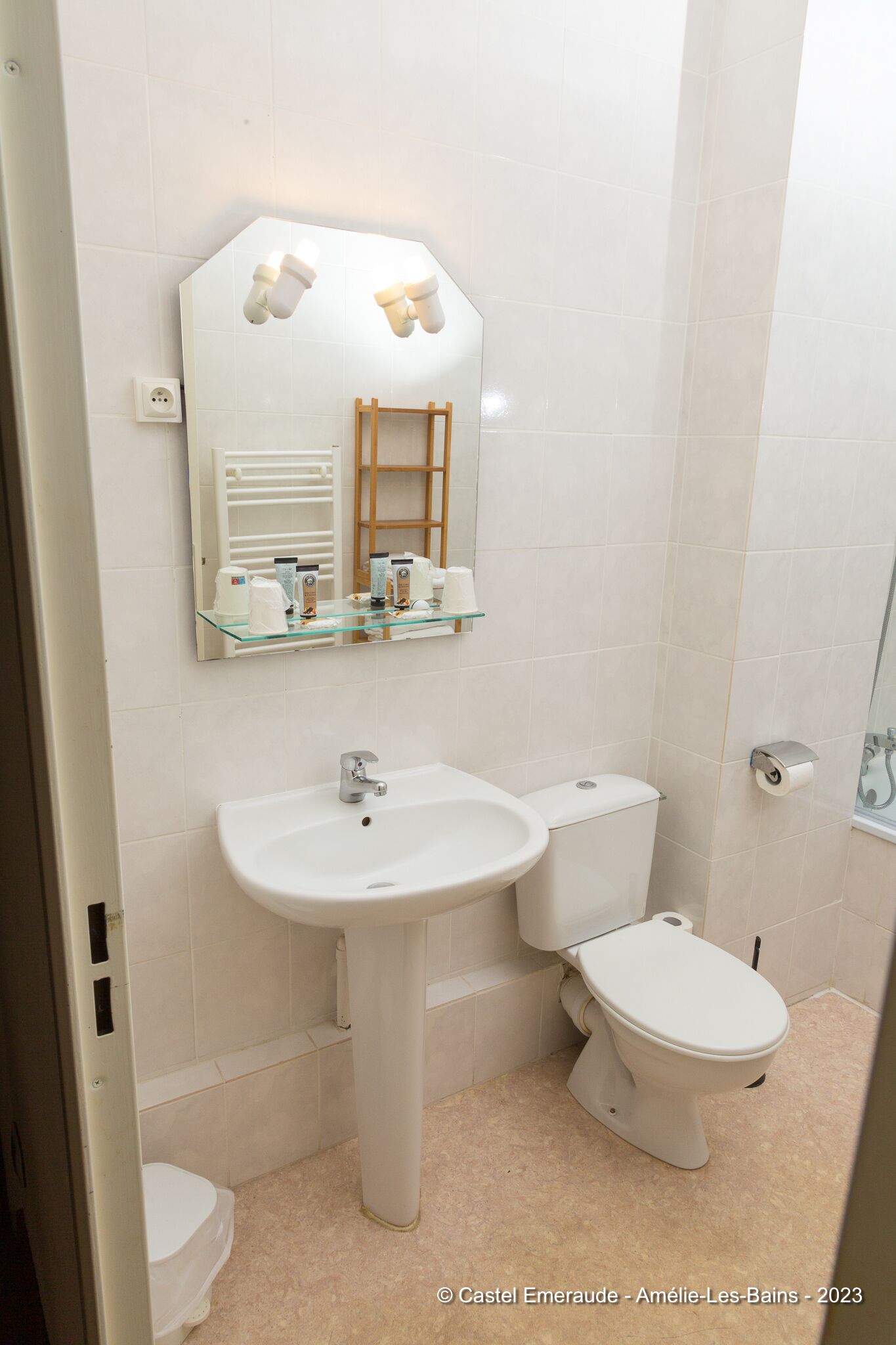 Un baño con lavamanos, inodoro y espejo blancos. Hay una repisa sobre el espejo con artículos de aseo.