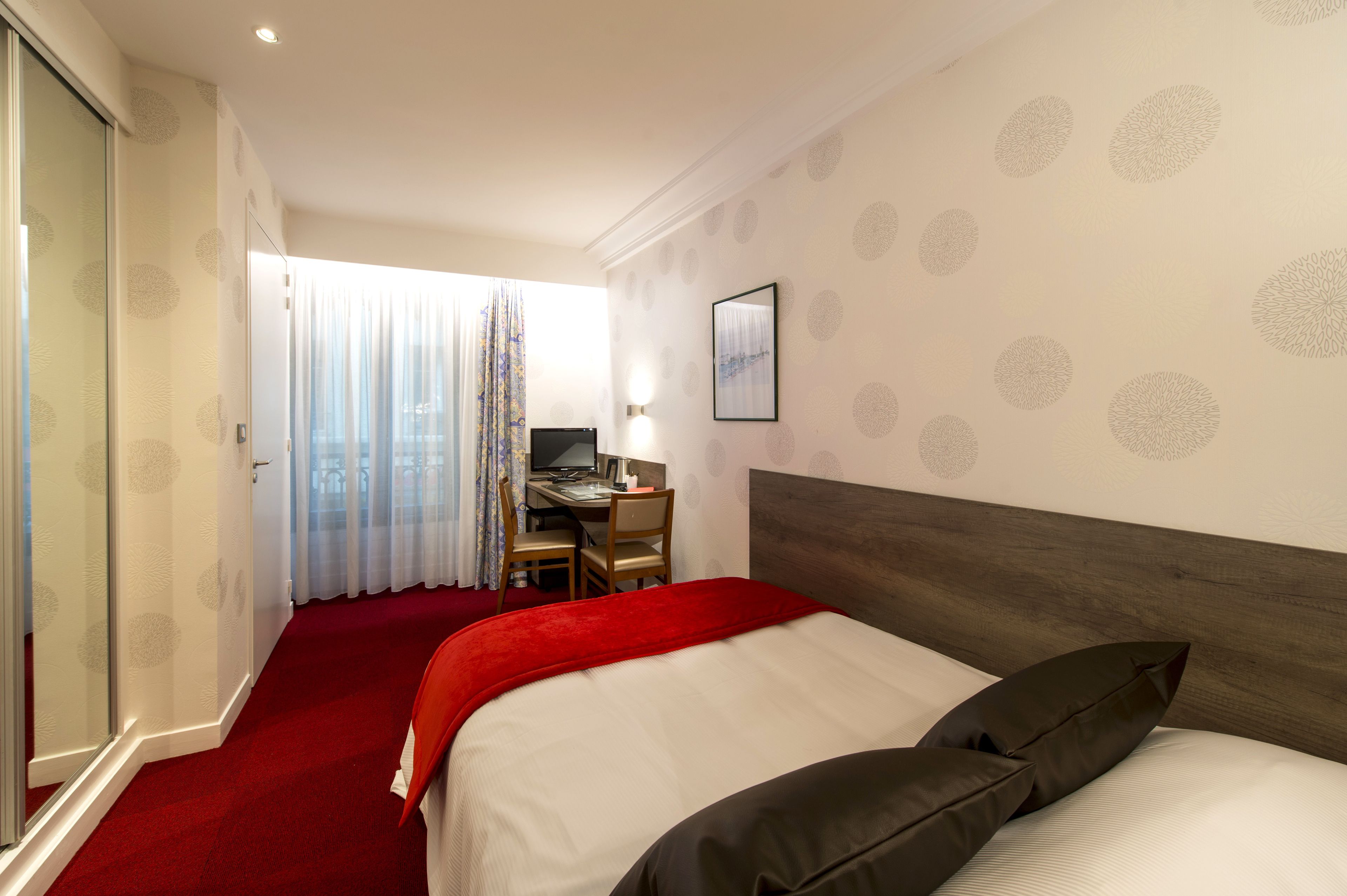 A hotel room with a bed, a desk with a computer, and a window with curtains.