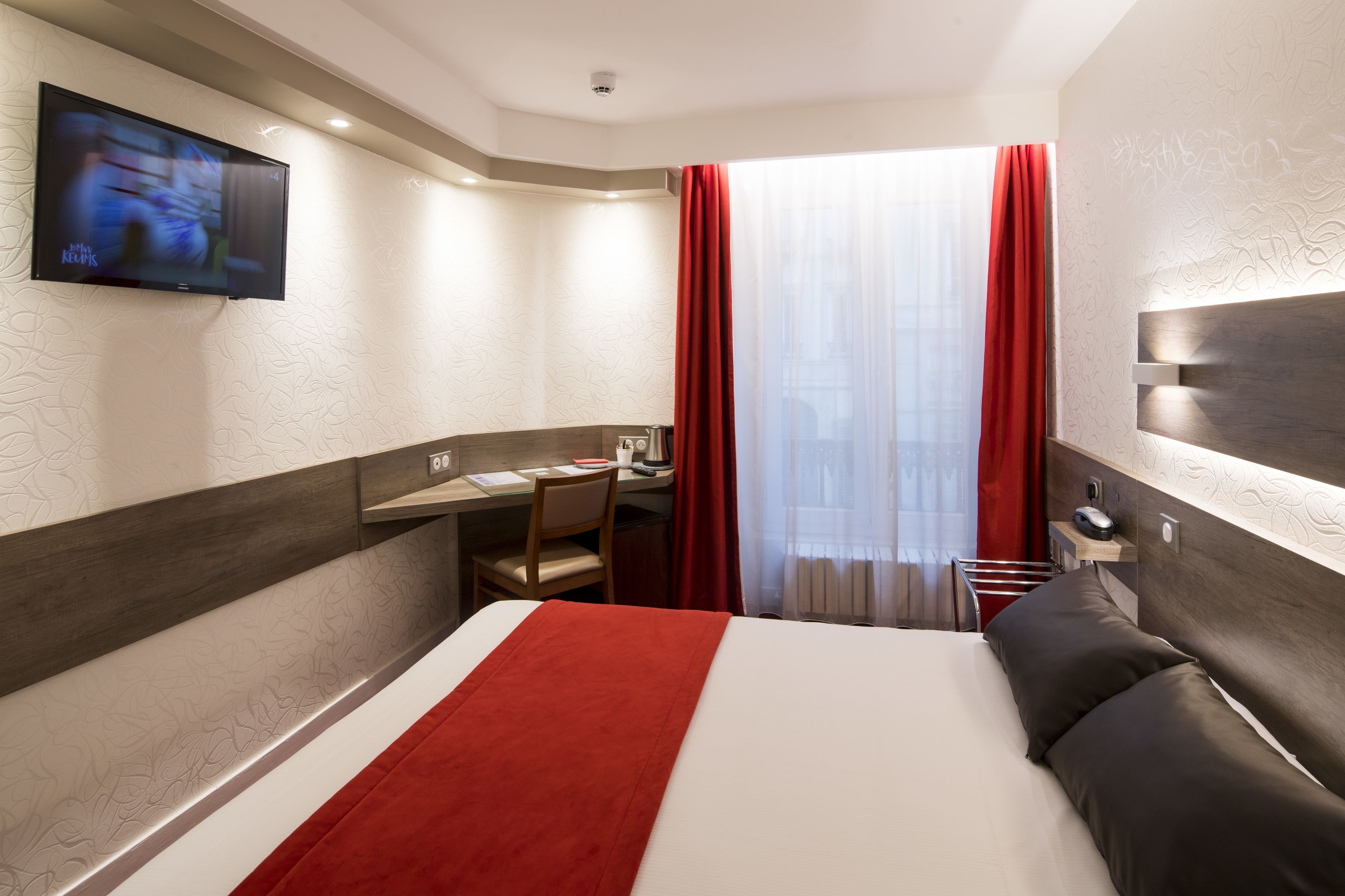 Een hotelkamer met een bed, een bureau met stoel, een aan de muur bevestigde televisie en een raam met rode gordijnen.
