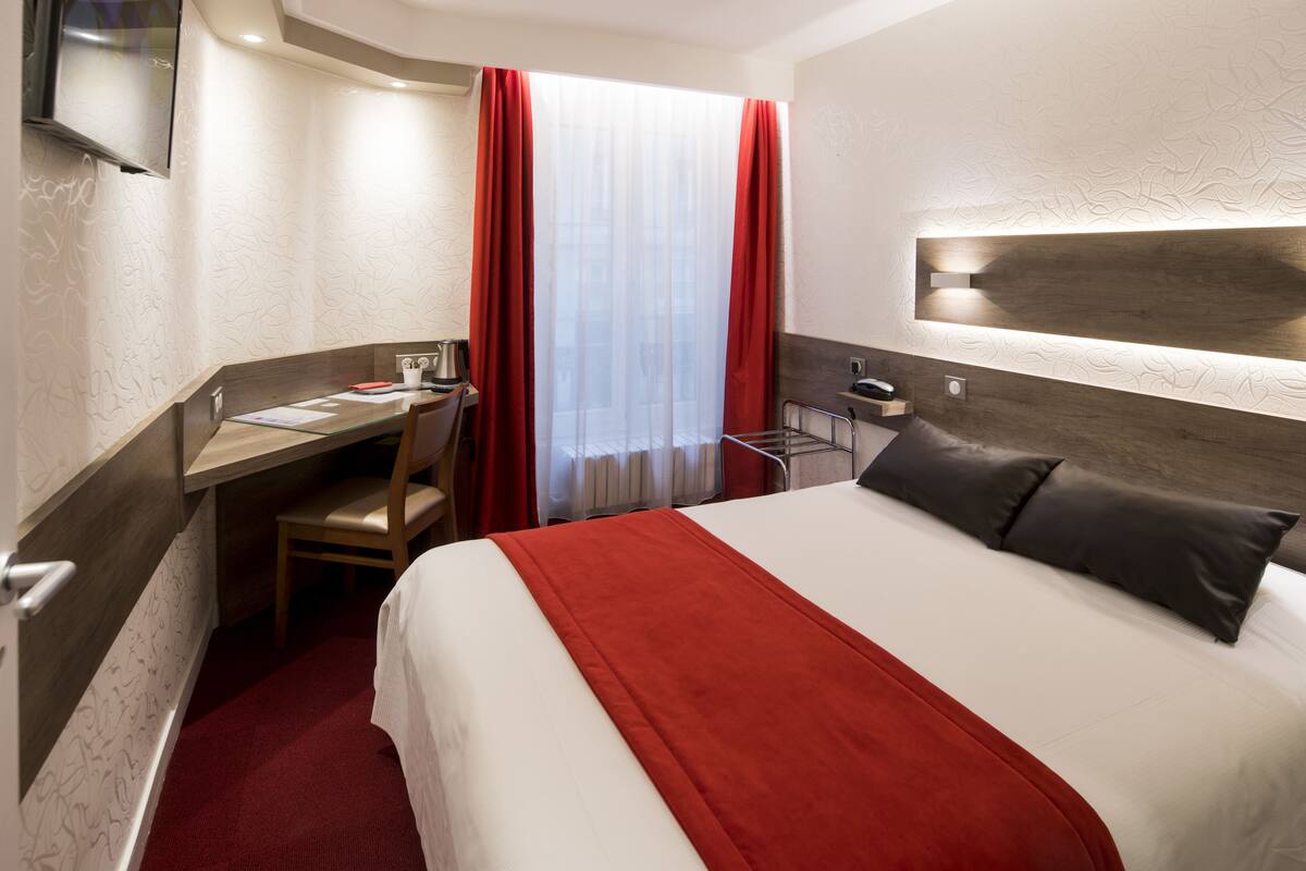 Habitación de hotel con cama, escritorio, silla, televisor y ventana con cortinas.