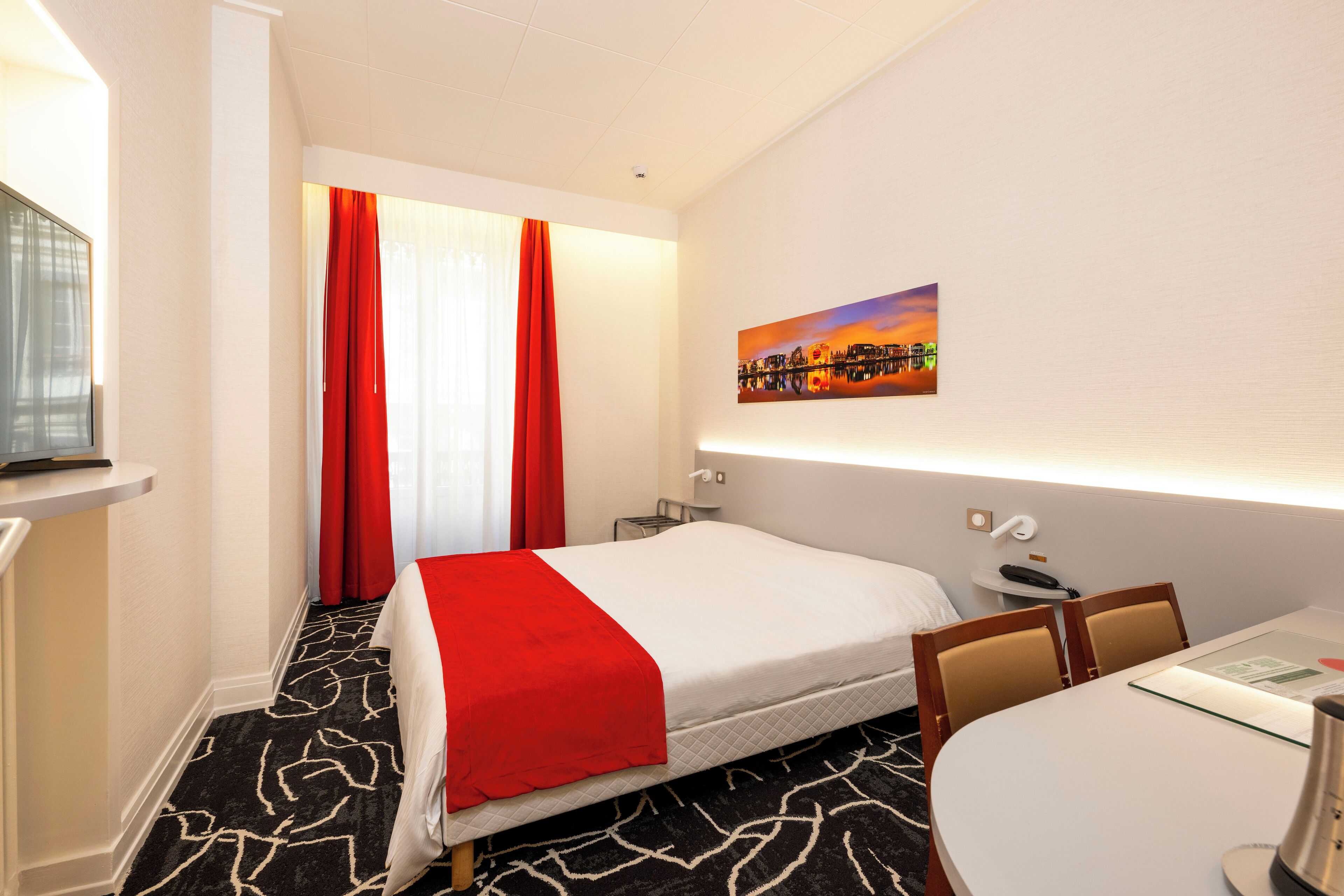 A hotel room with a bed, a desk, a chair, a painting on the wall, and a window with curtains.