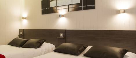 A hotel room with two beds, each with a red blanket, white bedding, and dark headboards. There are wall-mounted light fixtures and a framed artwork above the beds.