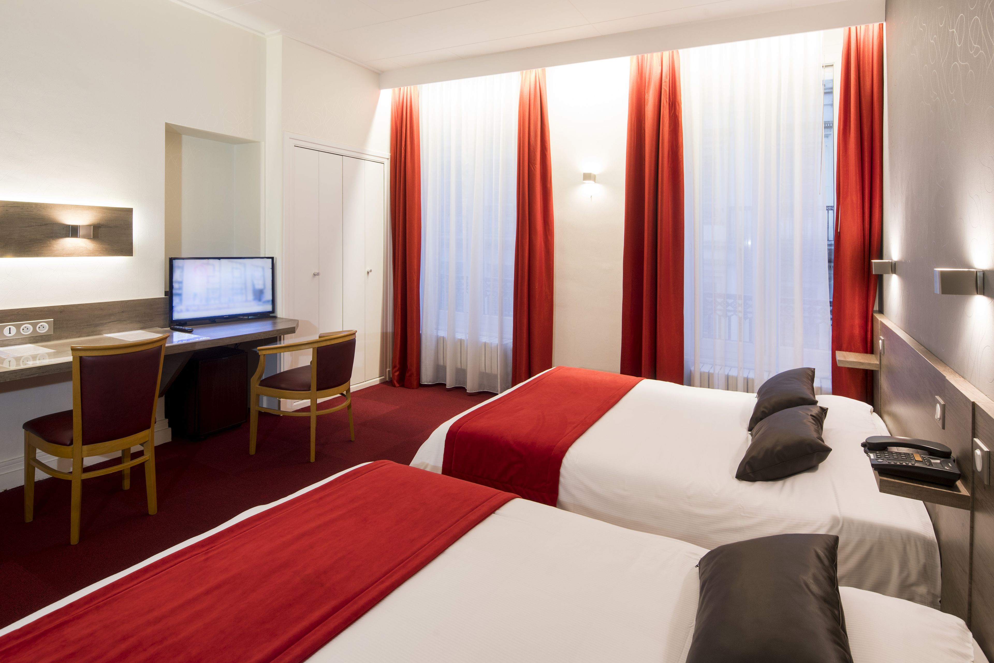 Een hotelkamer met twee bedden, een bureau met een computer en uitzicht op de stad door grote ramen.