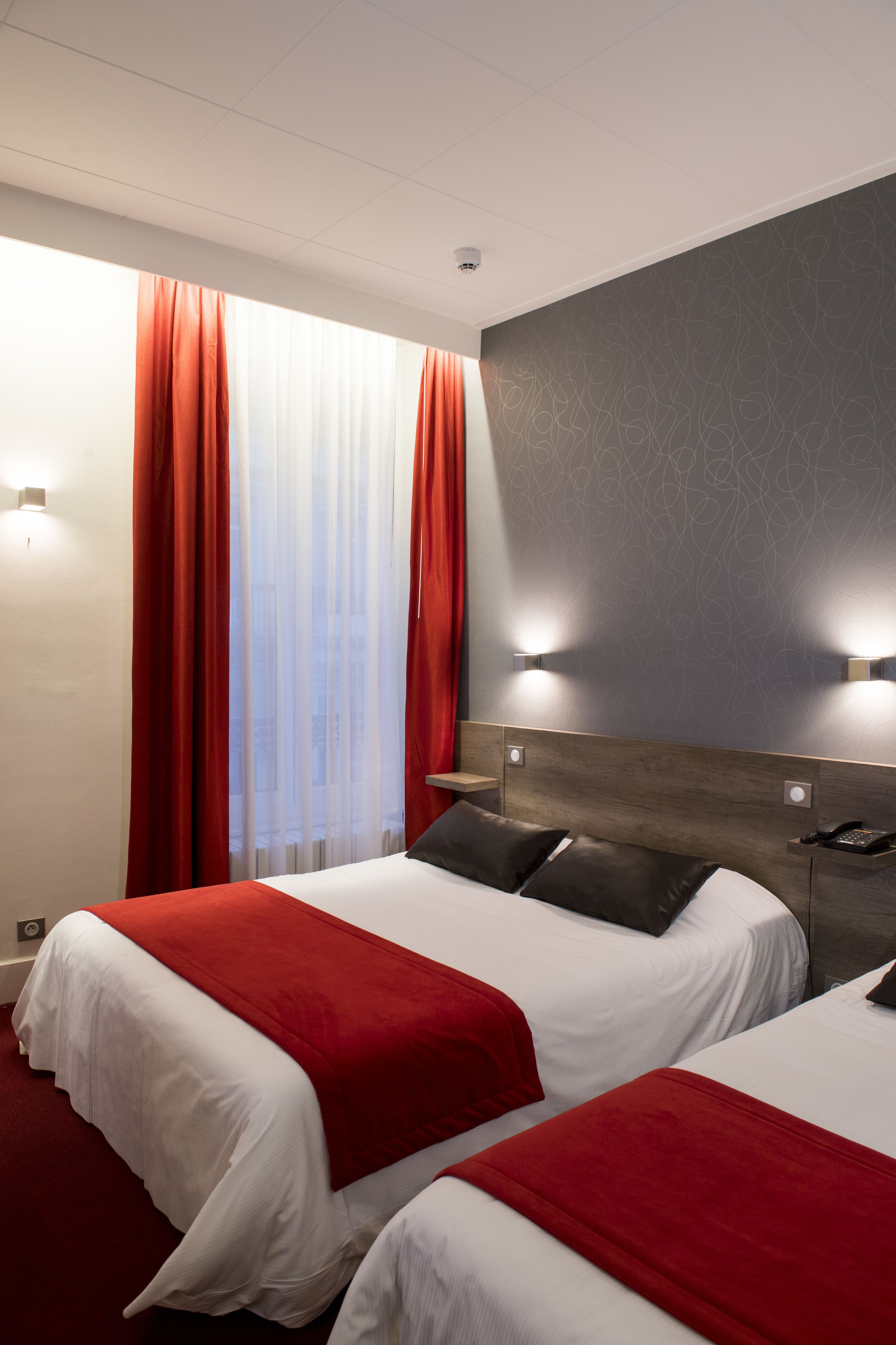 A hotel room with two beds, red and white bedding, a wooden headboard, and a window with red curtains.