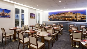 A hotel dining area with round tables and chairs, large windows, and cityscape paintings on the walls. - Hôtel La Résidence Lyon (Lyon)