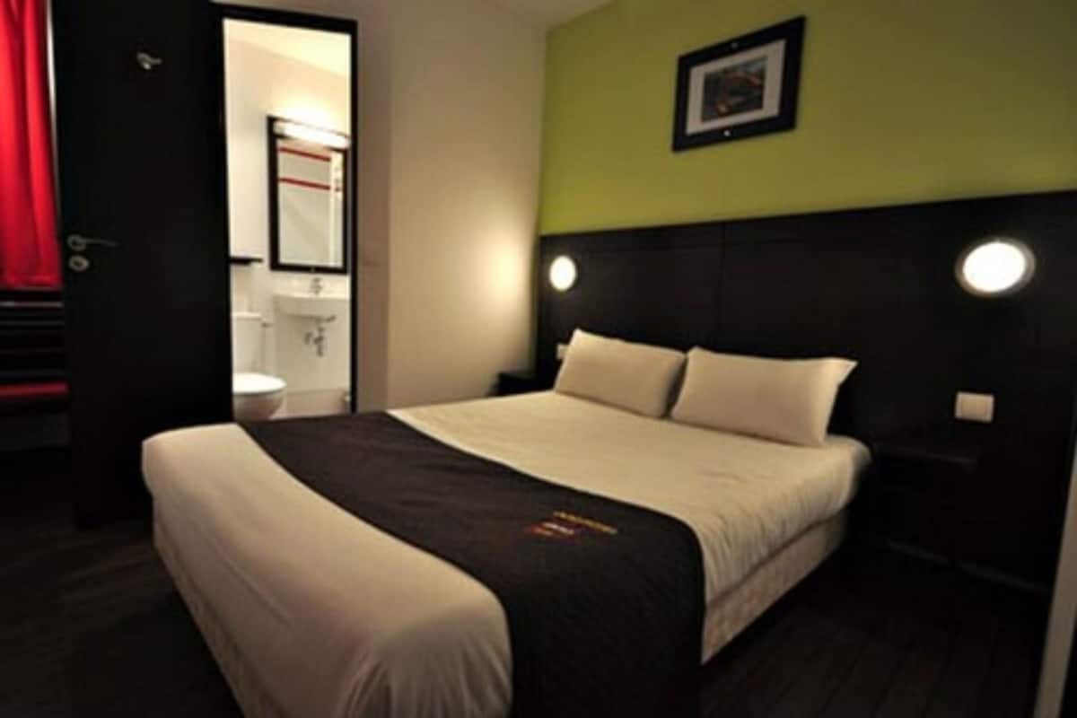 A hotel room with a bed, a nightstand, a wall-mounted light, and a door leading to a bathroom.
