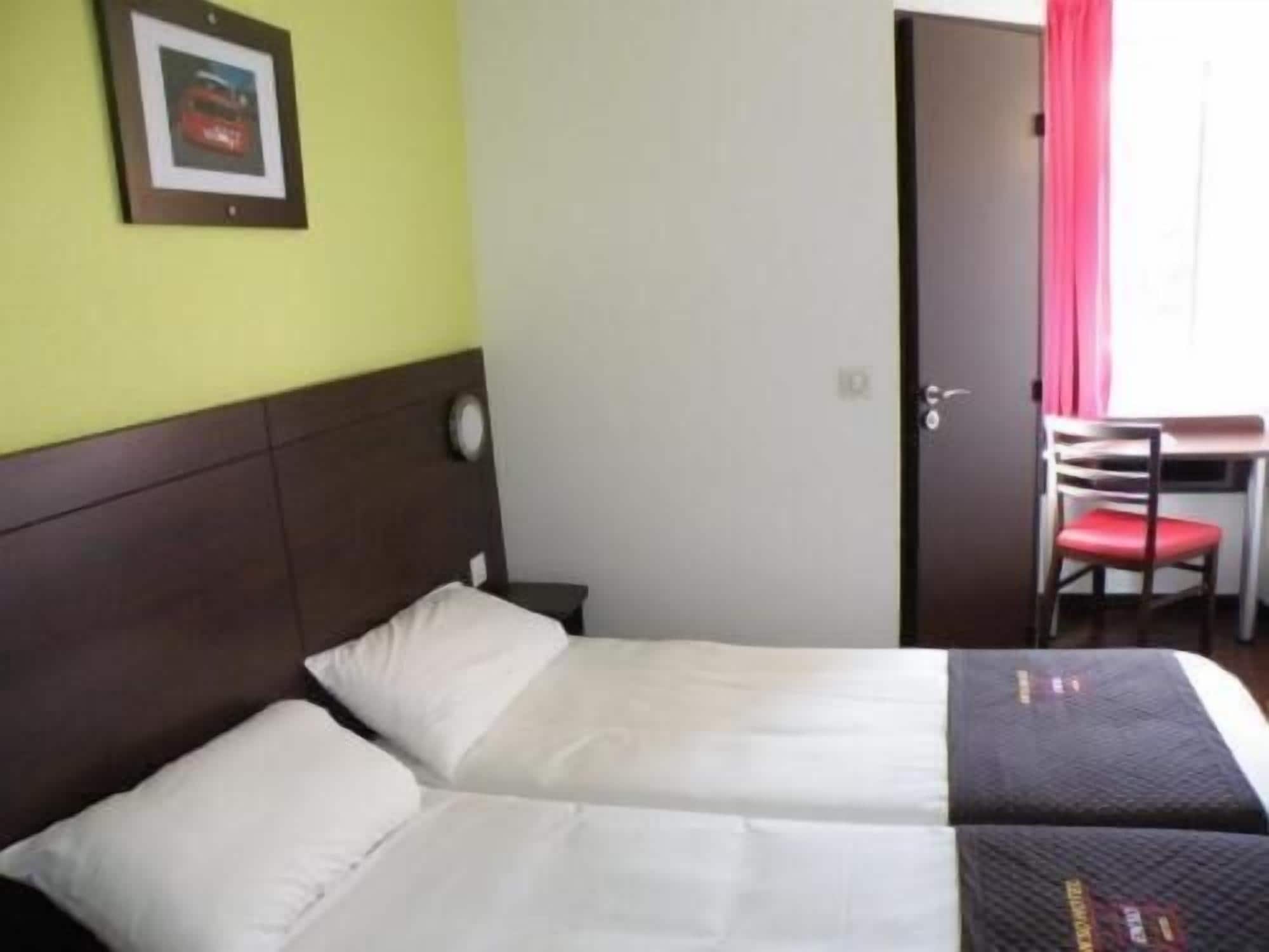 Un lit double avec du linge de lit blanc, une tête de lit en bois, une petite table de chevet, une chaise avec un coussin rouge et un tableau encadré au mur.