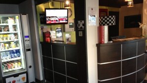 A reception area with a checkered floor mat, a vending machine, a television, and a counter with a checkered pattern. - Enzo Hotels Mulhouse Sud Morschwiller (Morschwiller-le-Bas)
