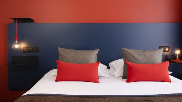 A bed with white linens, two red pillows, and a blue headboard. - Hôtel Le Bugatti (Molsheim)