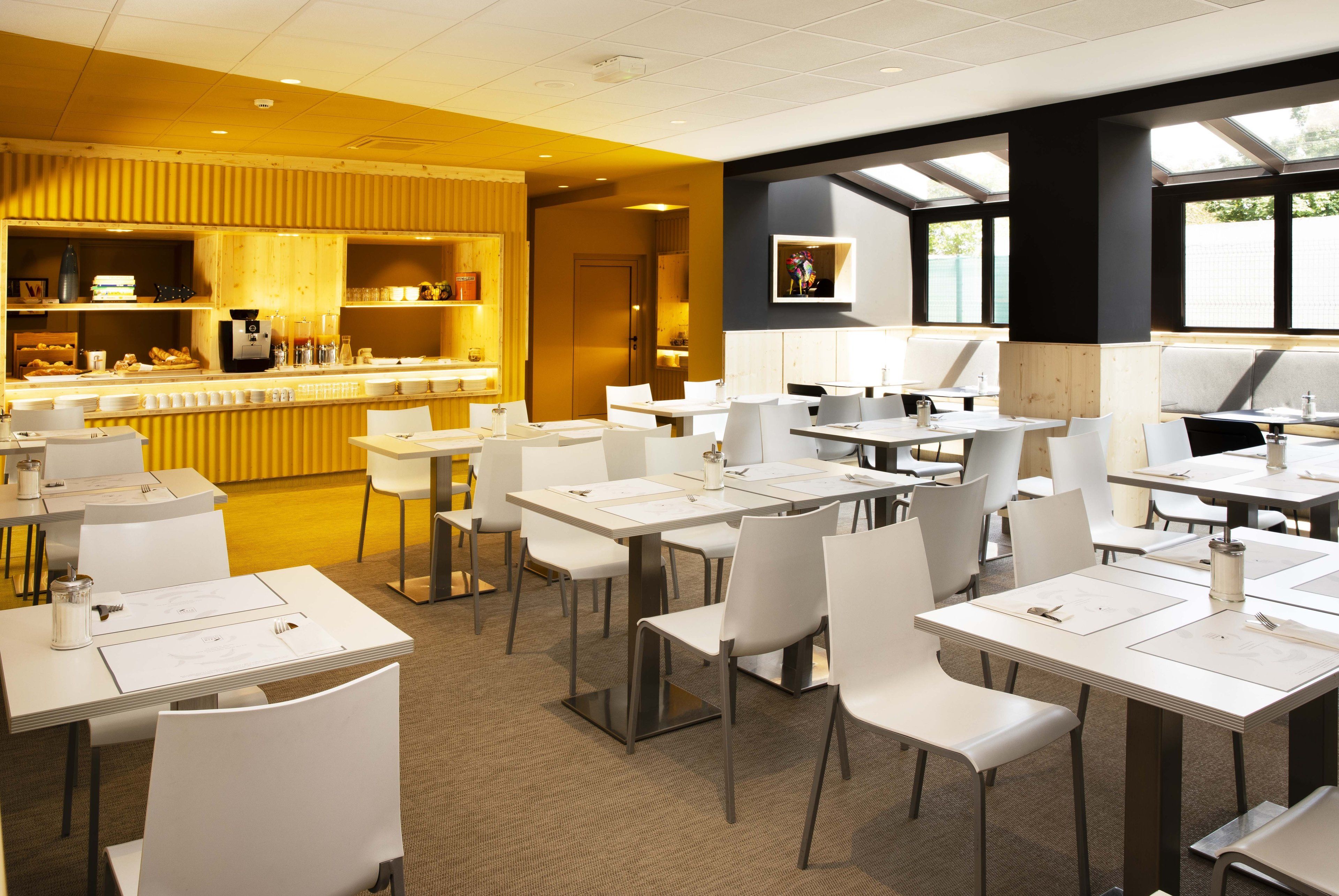 A modern restaurant with white tables and chairs, a bar area, and large windows providing natural light.