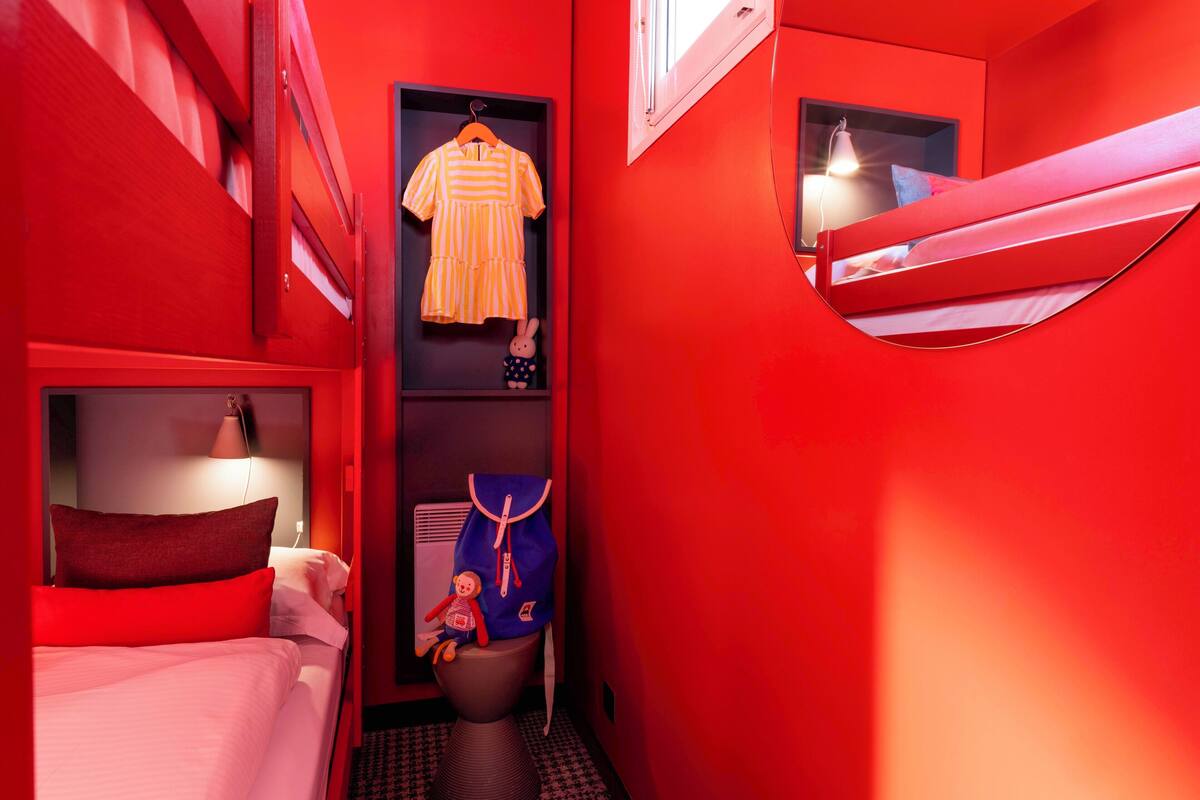 A small, red-themed room with a bed, a hanging wardrobe, a small shelf with a doll, and a window.