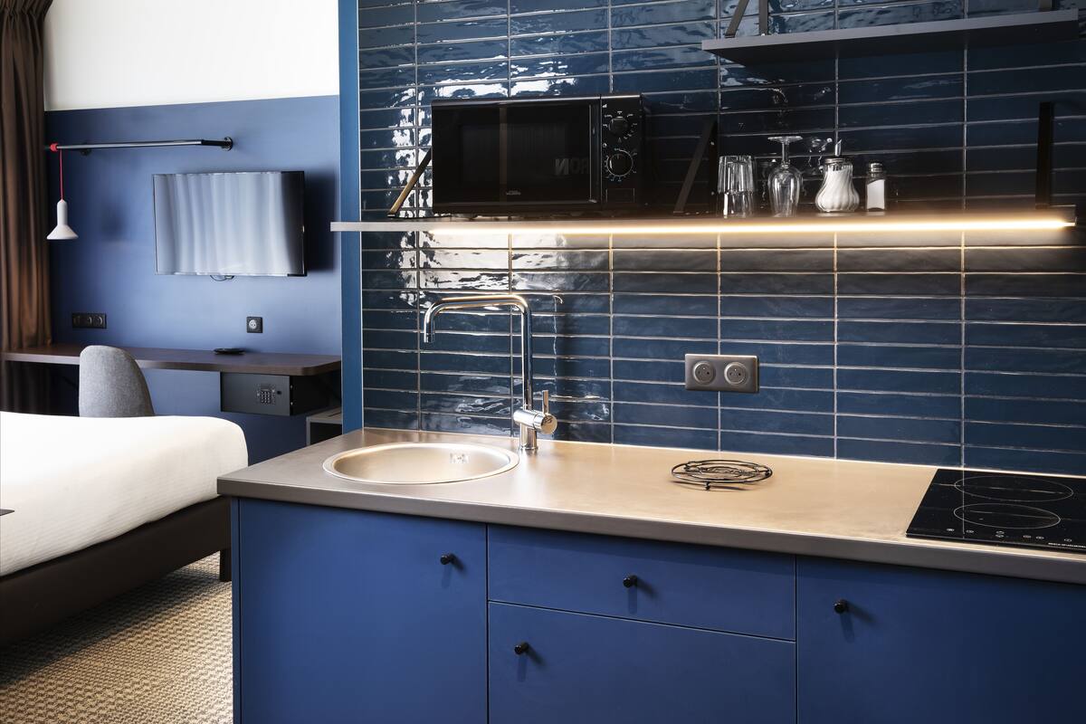 A modern hotel room with a bed, a desk, a flat-screen TV, a microwave, a sink, and a built-in stove.