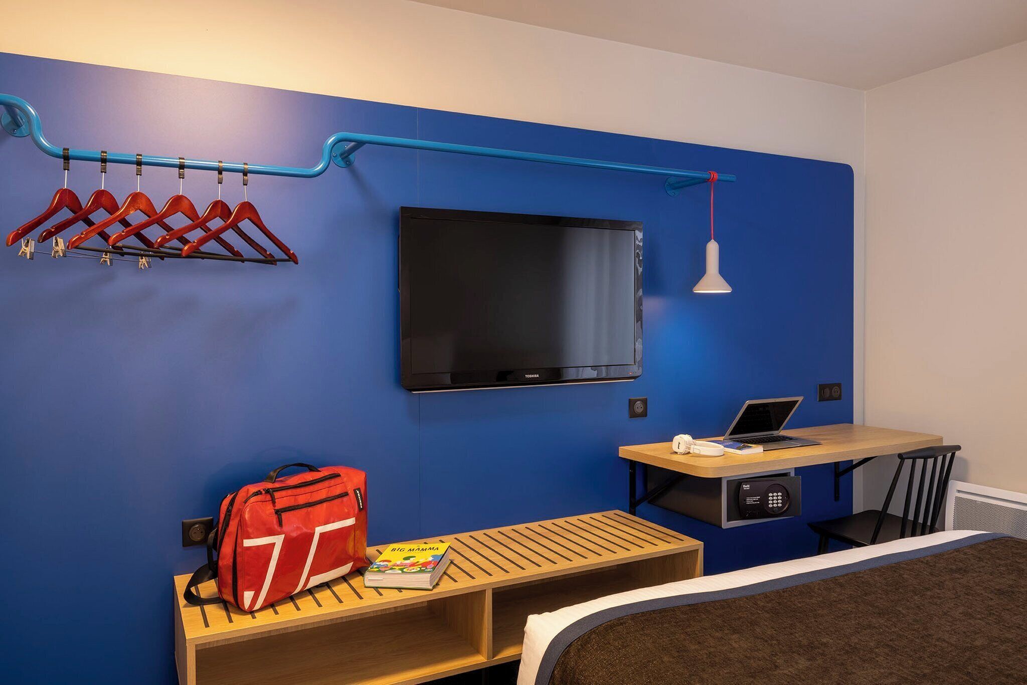 Habitación de hotel con pared azul, televisor montado, escritorio con una computadora portátil, cama y un banco con un bolso rojo y un libro.