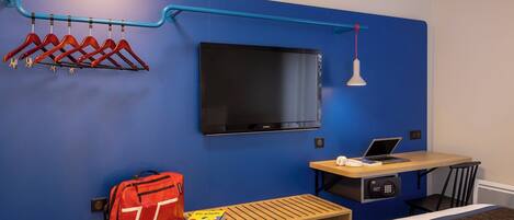 A hotel room with a blue wall, a mounted TV, a desk with a laptop, a bed, and a bench with a red bag and a book.