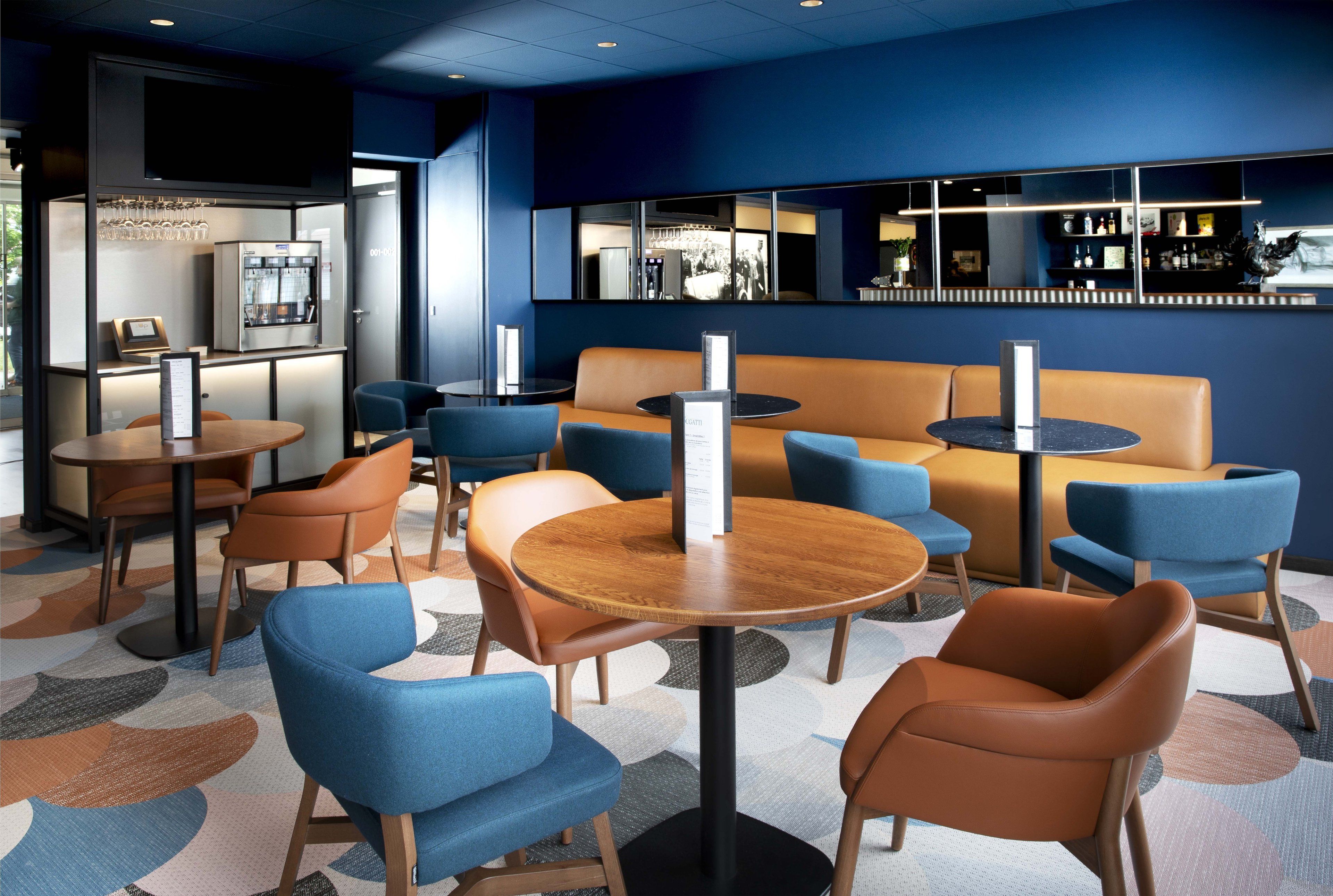 Un moderno vestíbulo de hotel con mesas redondas, sillas azules y marrones, una zona de bar y un área de estar con sofá y sillones.