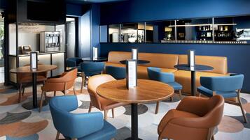 Un moderno vestíbulo de hotel con mesas redondas, sillas azules y marrones, una zona de bar y un área de estar con sofá y sillones.