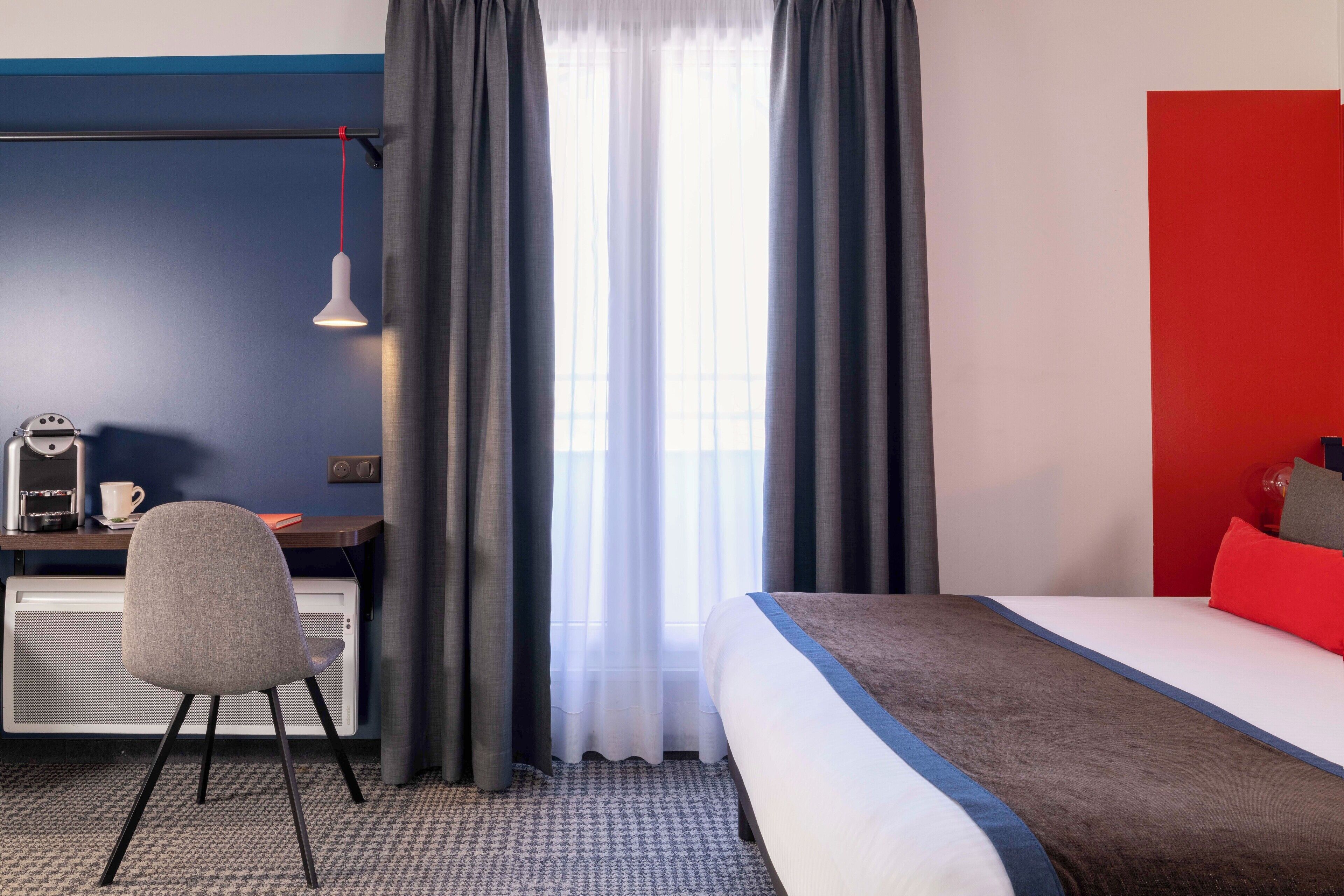 A hotel room with a bed, a desk with a chair, a radiator, and a window with curtains.