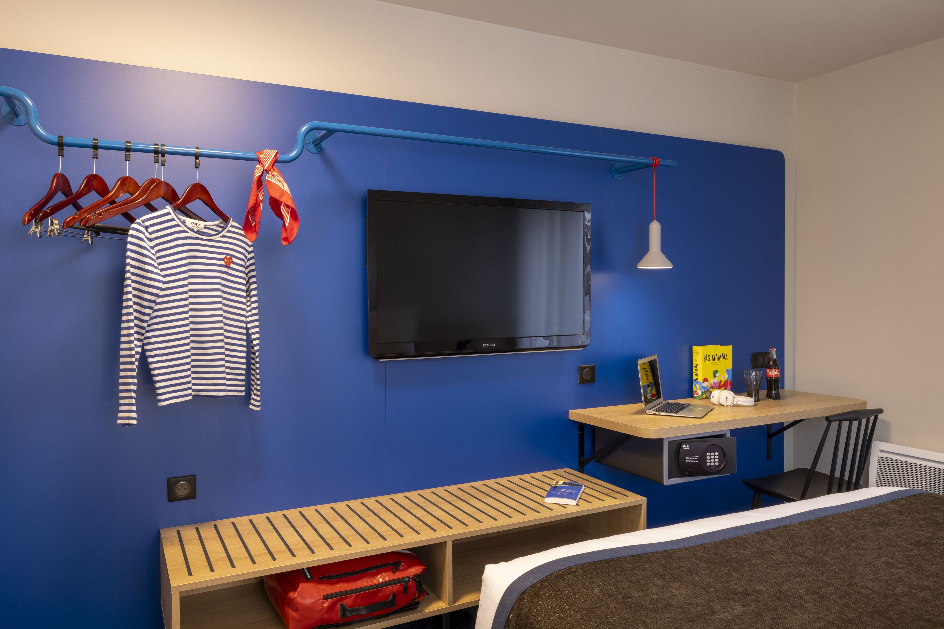 Une chambre d’hôtel avec un mur bleu, un téléviseur fixé au-dessus d’un banc en bois, un bureau avec un ordinateur portable et un lit doté d’une tête de lit marron.