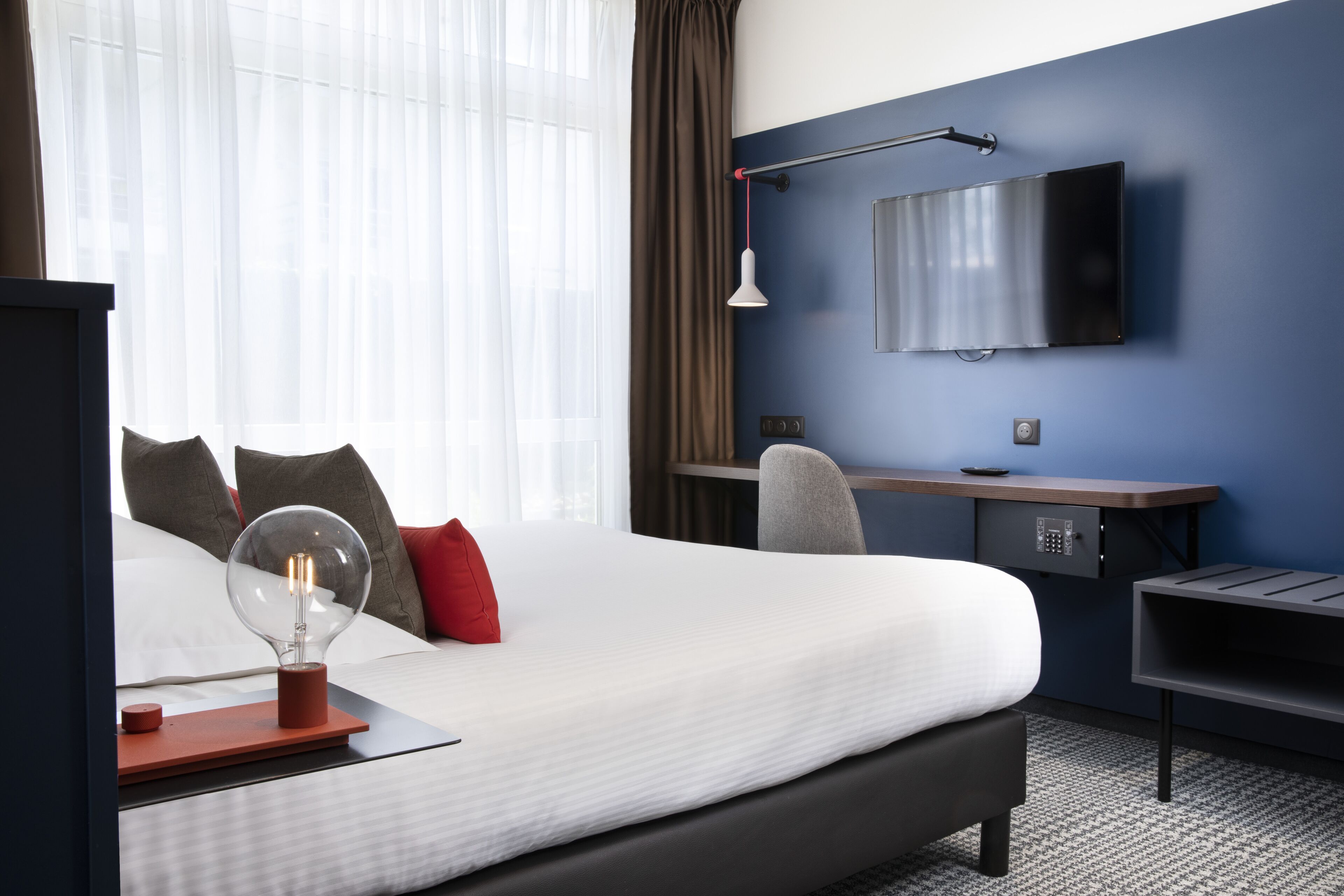 A hotel room with a large bed, a desk, a chair, a TV mounted on the wall, and a lamp.