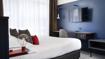 A hotel room with a large bed, a desk, a chair, a TV mounted on the wall, and a lamp.