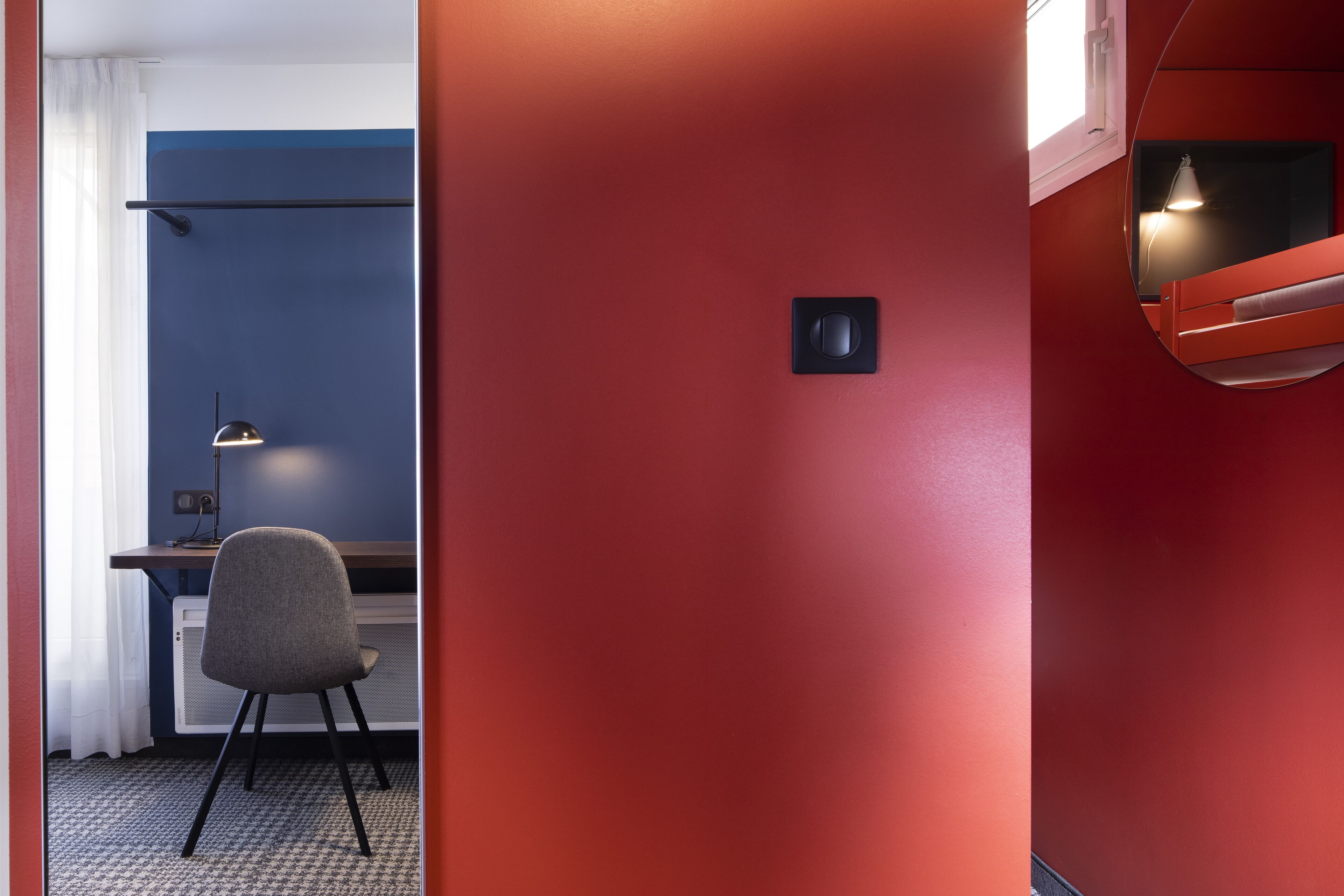 A room with a red wall, a desk with a chair, and a bed with a headboard.