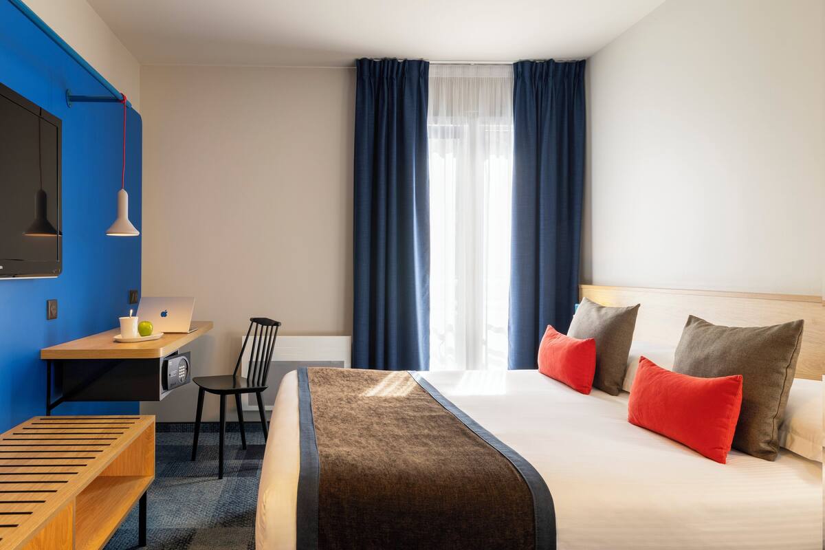 A hotel room with a bed, a desk with a laptop, a chair, and a window with curtains.