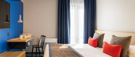 A hotel room with a bed, a desk with a laptop, a chair, and a window with curtains.