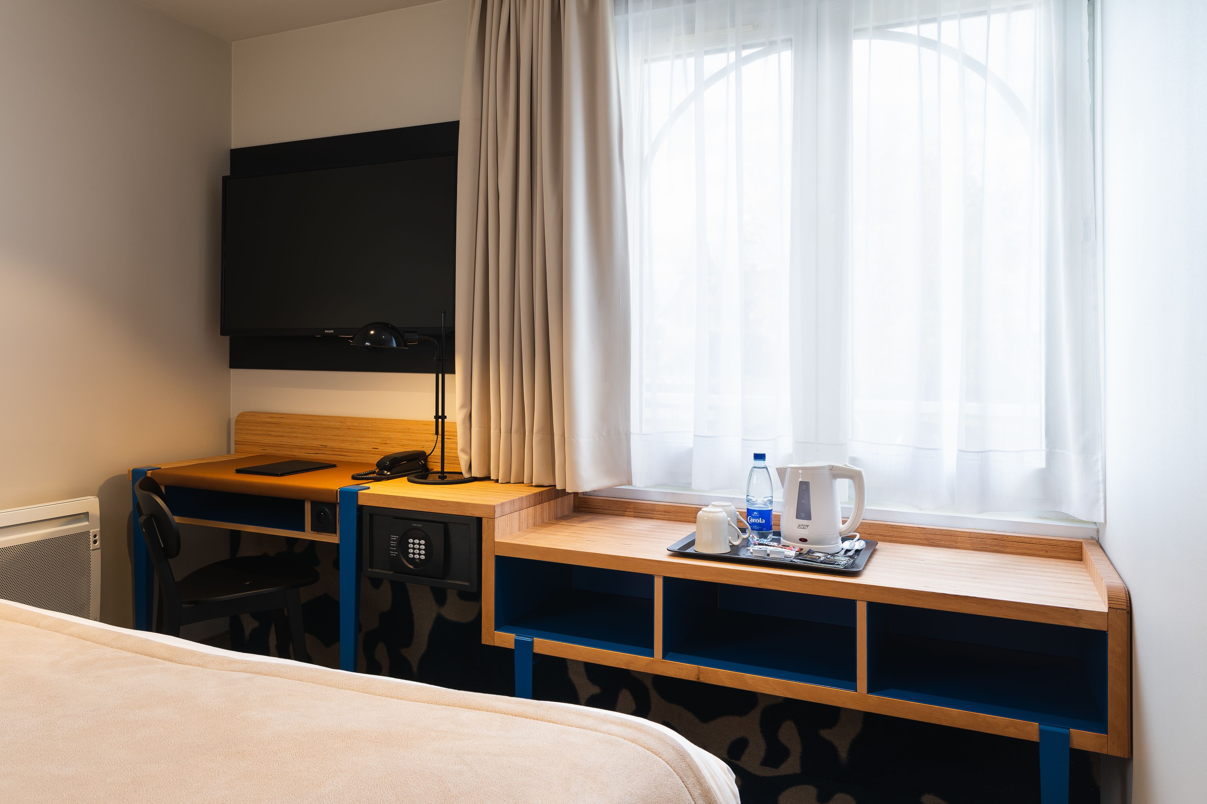 A hotel room with a bed, a desk with a computer, a flat-screen TV, a window with curtains, and a coffee maker.
