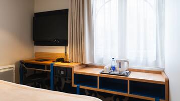 A hotel room with a bed, a desk with a computer, a flat-screen TV, a window with curtains, and a coffee maker.