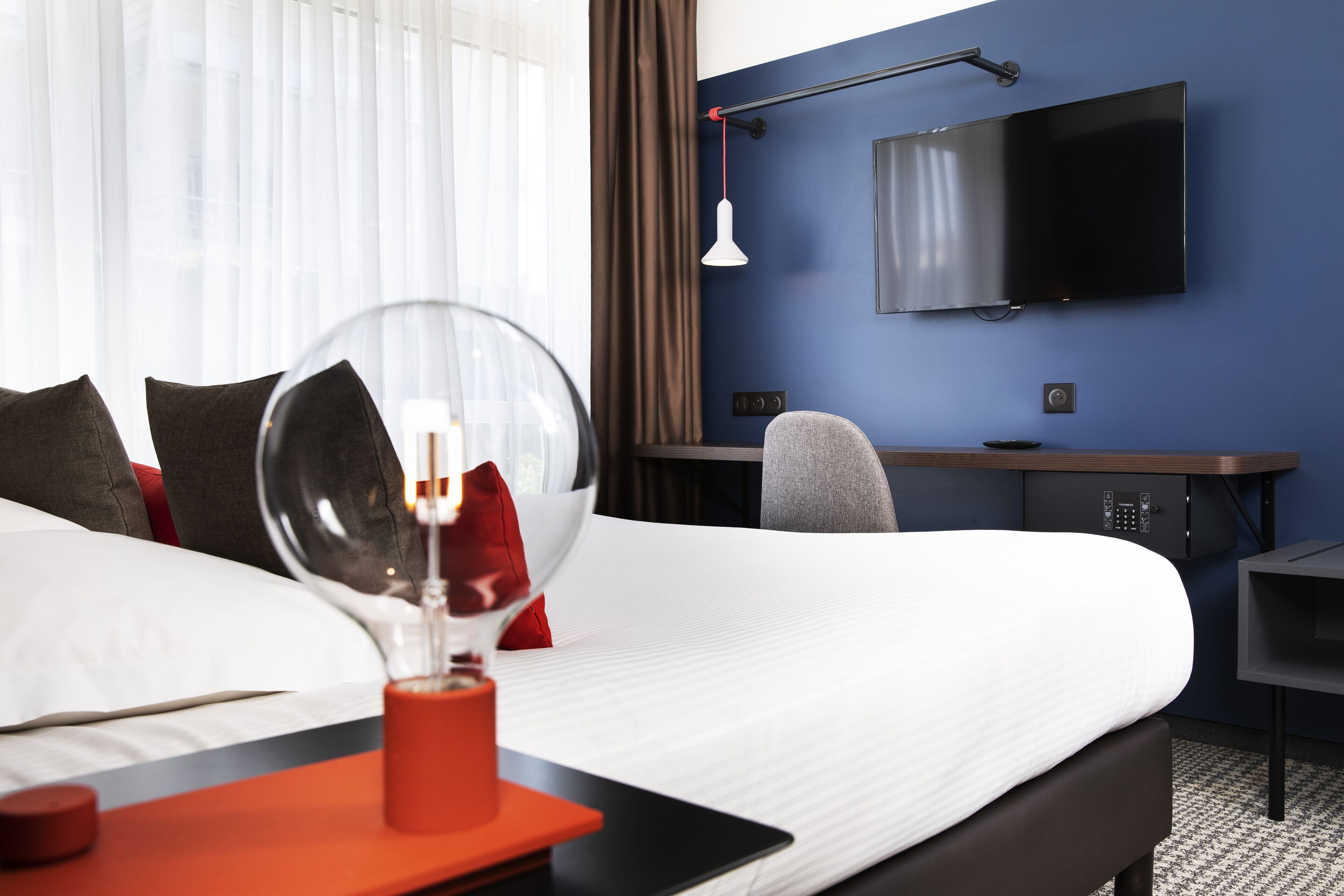 A hotel room with a large bed, a desk with a chair, a TV mounted on the wall, and a bedside table with a lamp.
