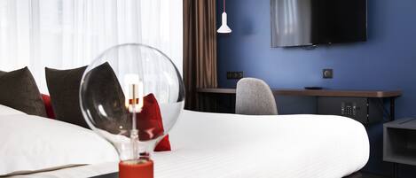 A hotel room with a large bed, a desk with a chair, a TV mounted on the wall, and a bedside table with a lamp.