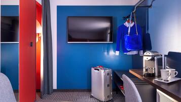 Habitación de hotel con televisión, escritorio con cafetera, maleta y bolso colgado.