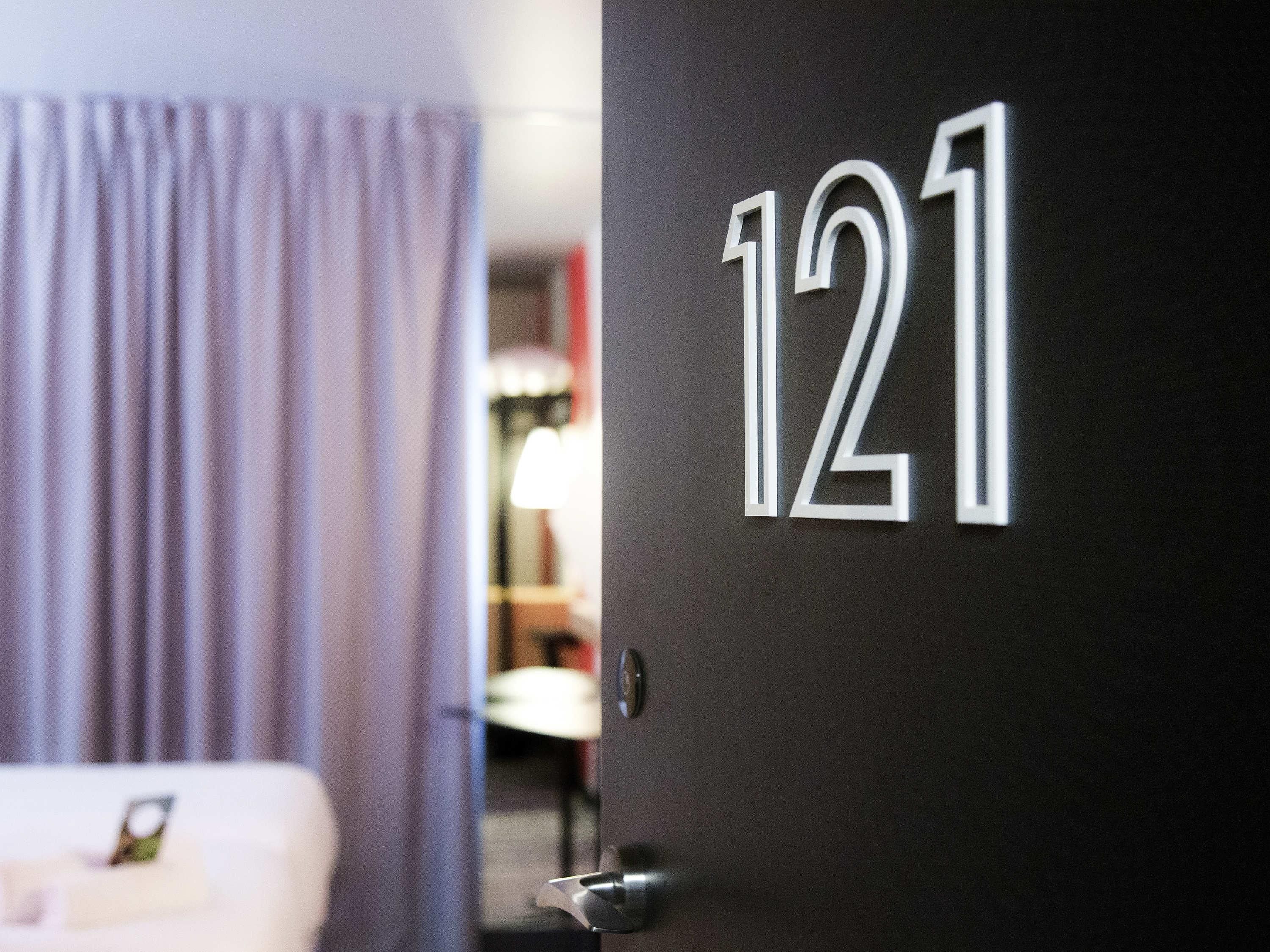 Een deur met het nummer 121, een kamer met een bed en een lamp, en een gedeeltelijk zichtbaar gordijn.