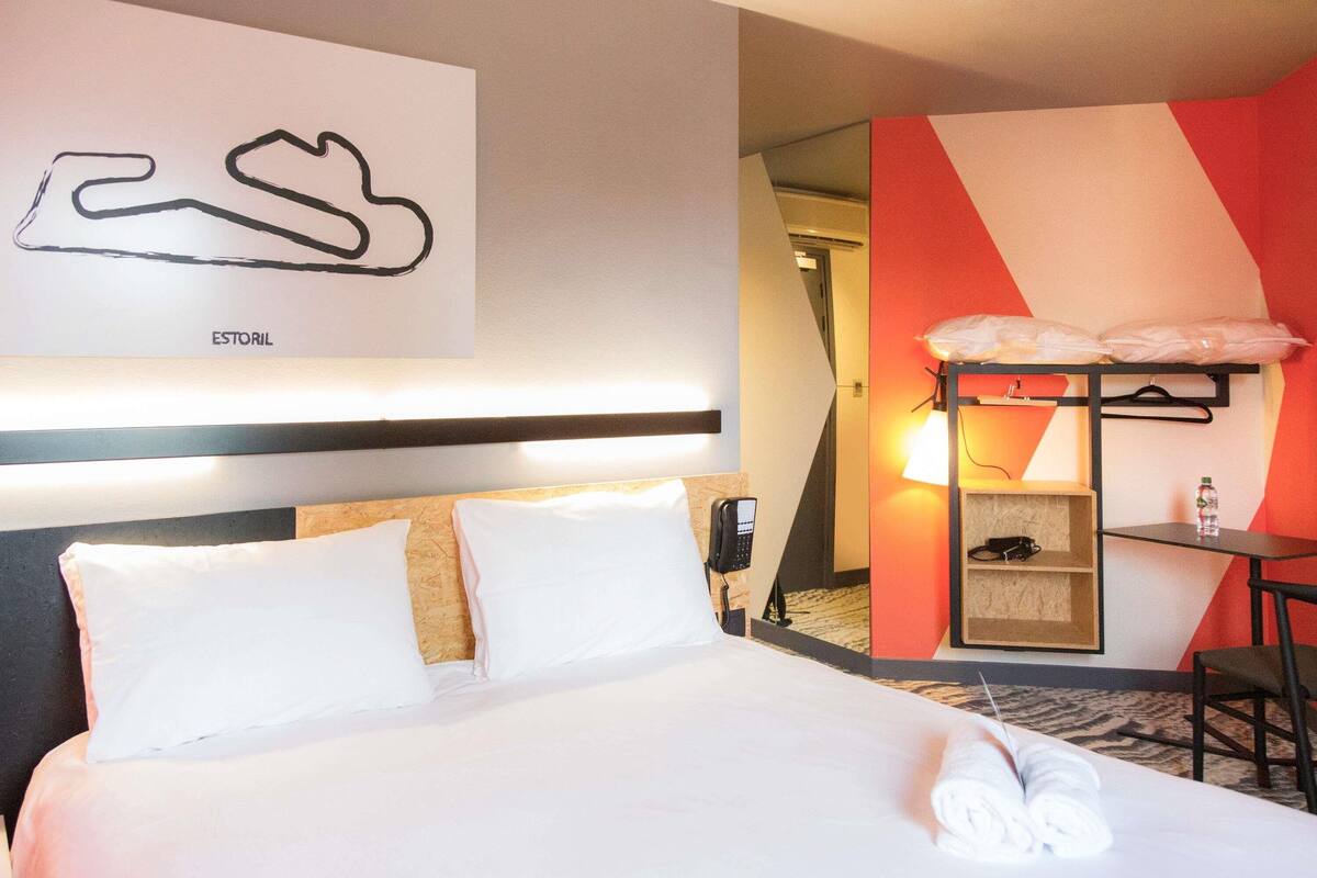 A hotel room with a bed, a desk with a chair, a phone, and a wall-mounted shelf with a lamp.