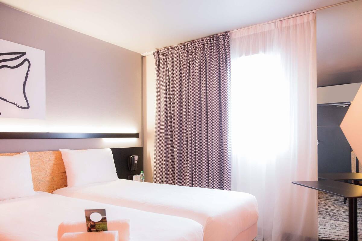 A hotel room with two beds, a bedside table, a lamp, and a large window with curtains.
