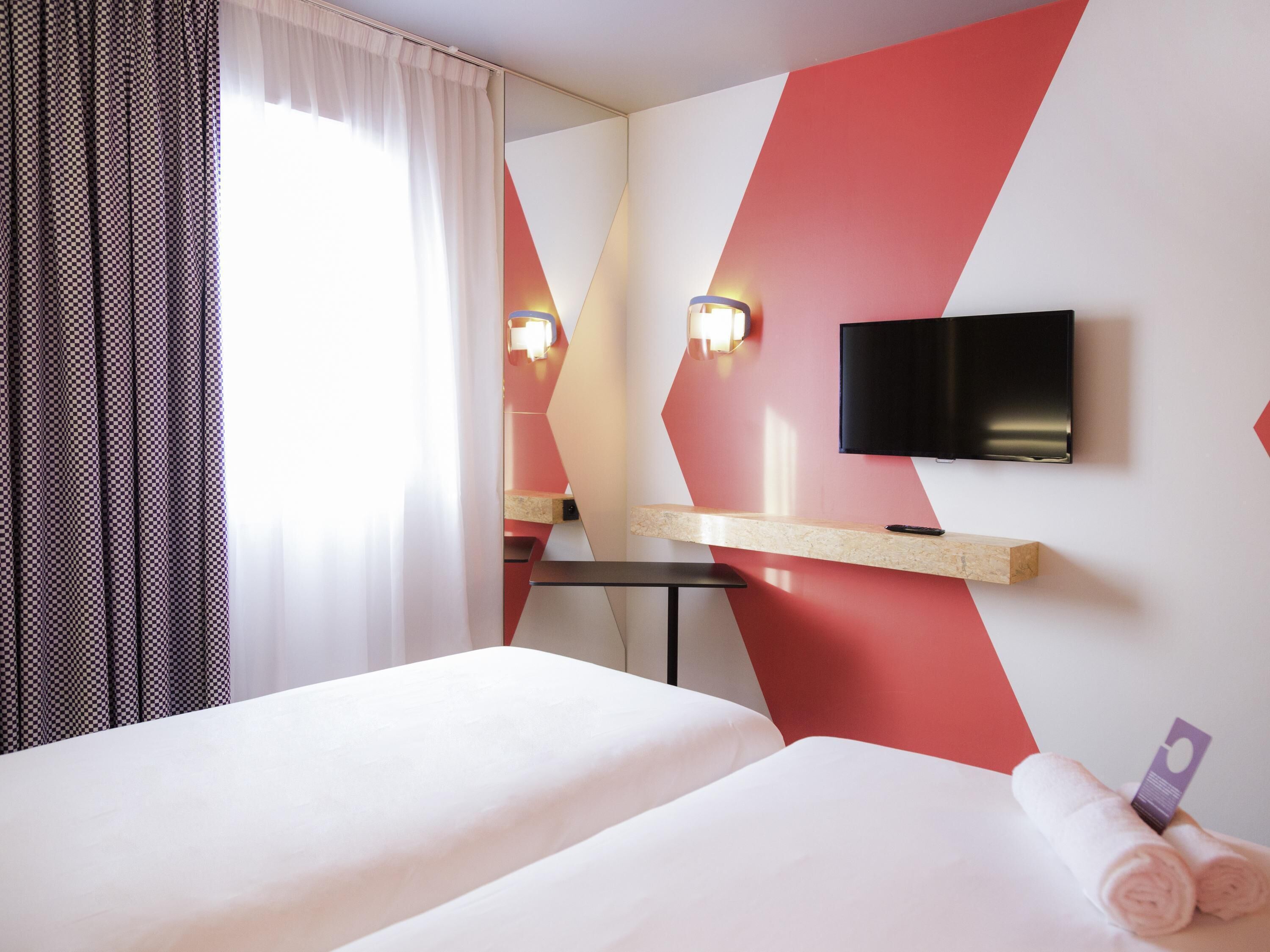 A hotel room with two beds, a TV, a desk, and a window with curtains.