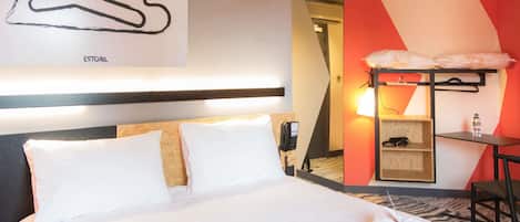 A hotel room with a bed, a desk with a chair, a phone, and a wall-mounted shelf with a lamp.