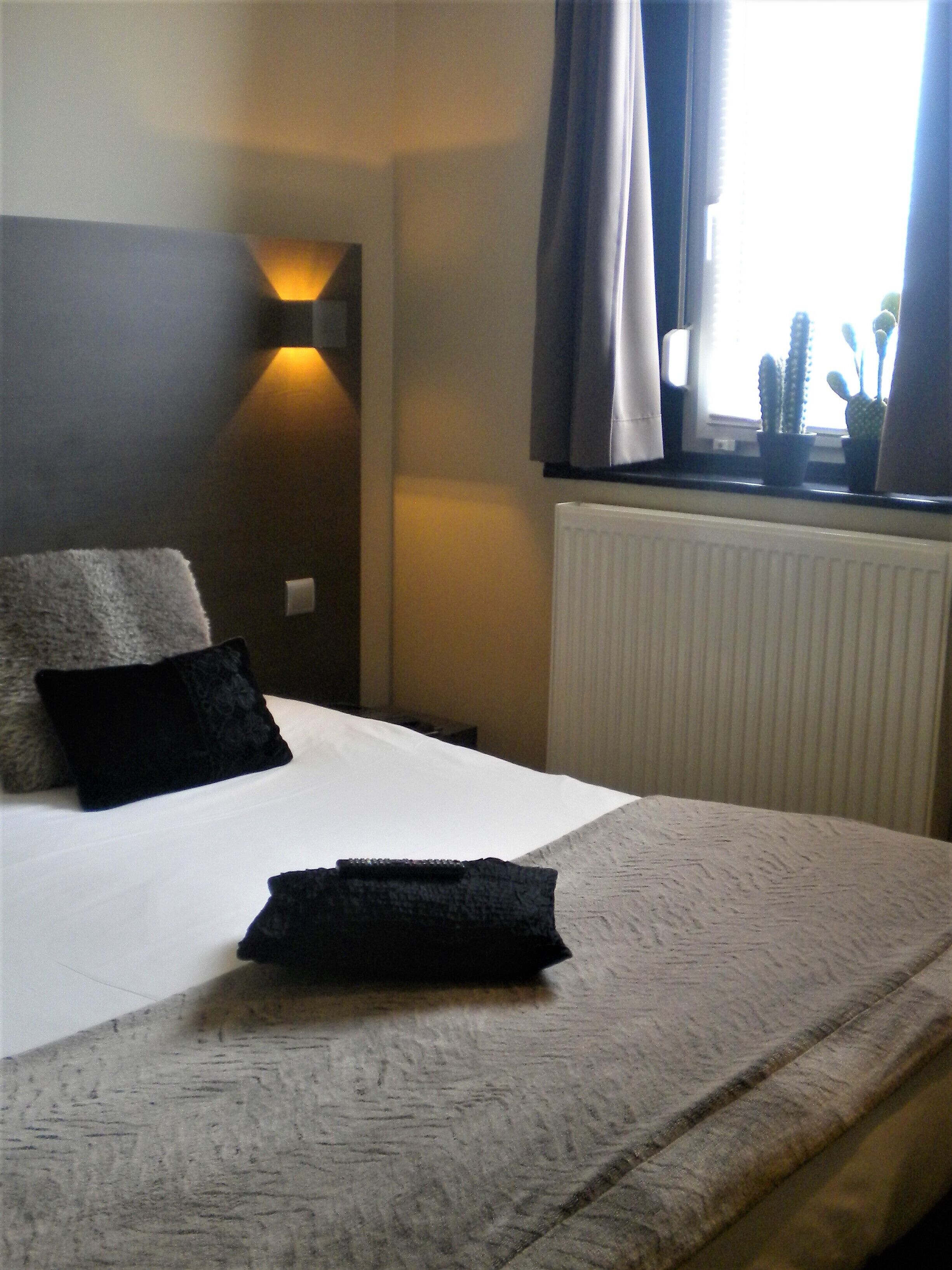 Een hotelkamer met een bed, nachtkastje, een wandlamp, gordijnen voor het raam en een radiator.