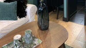 Une pièce moderne aux murs de couleur turquoise, agrémentée d’un miroir rond, d’un vase avec des fleurs blanches, d’une table basse en bois et d’une petite table d’appoint sur laquelle reposent deux tasses.