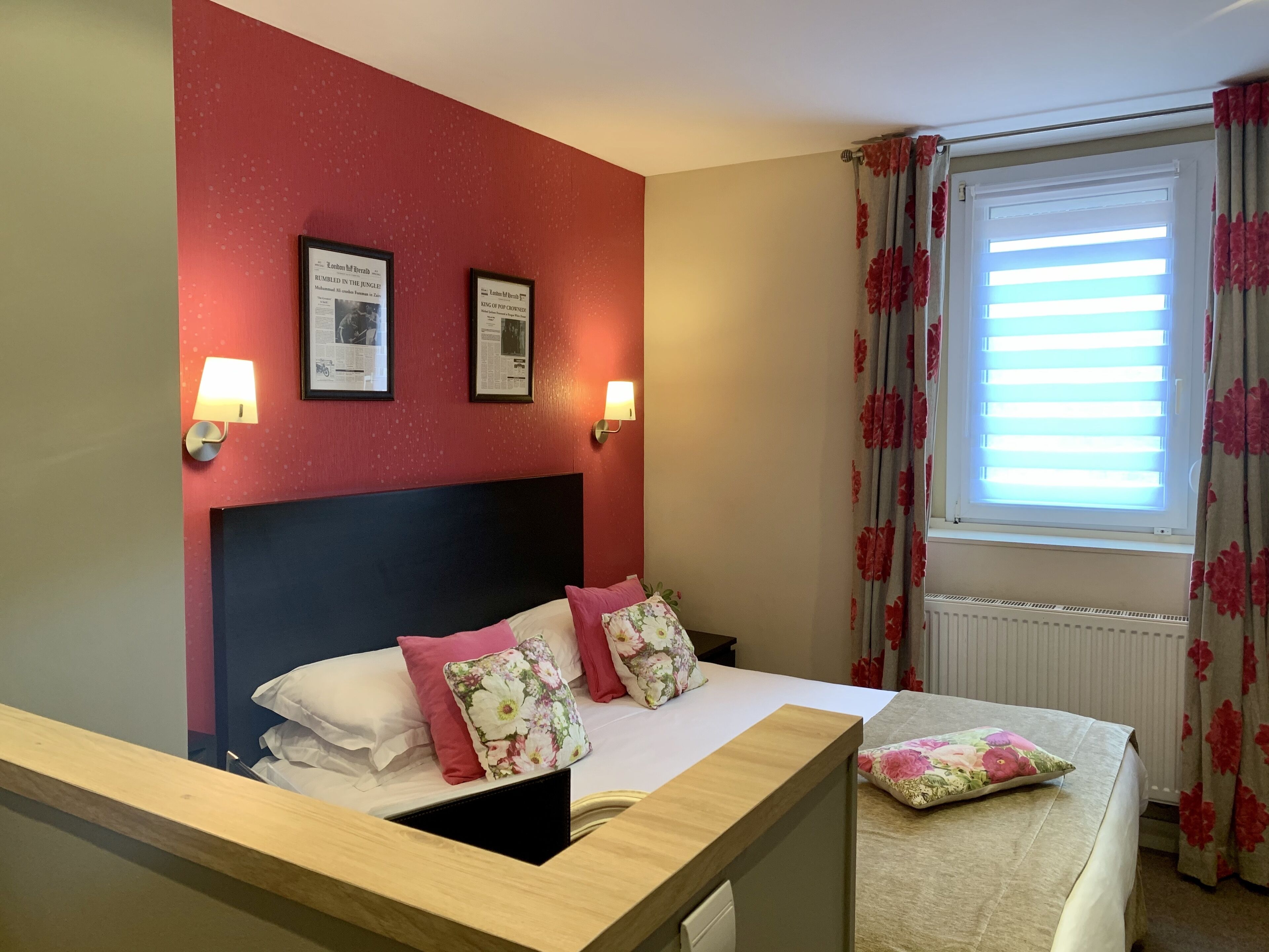 A hotel room with a bed, two wall-mounted lamps, a window with floral curtains, and two framed pictures on the wall.