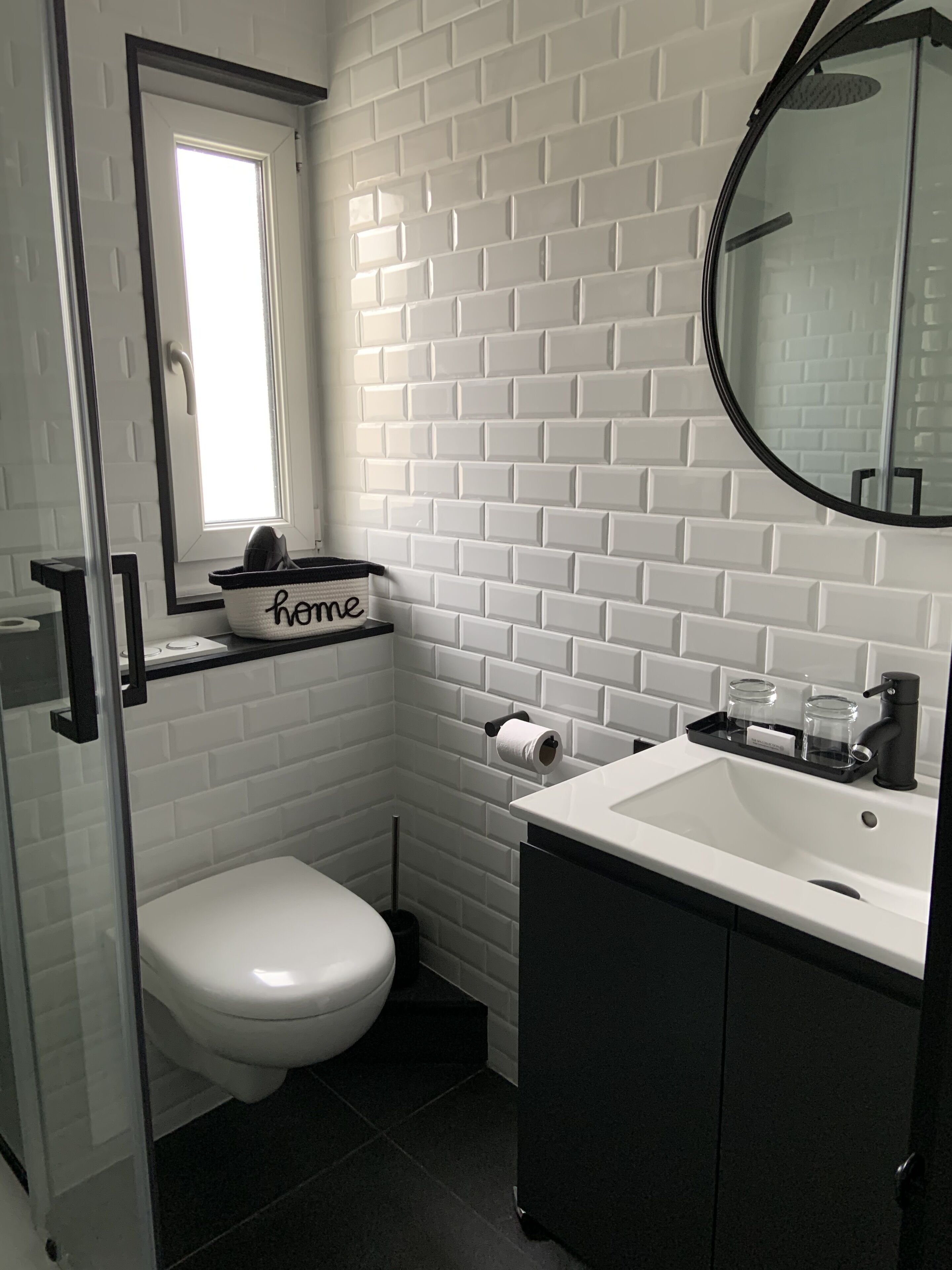 Un baño pequeño con azulejos blancos tipo metro, un inodoro blanco, un lavamanos negro con un espejo encima y una ventana pequeña con vistas al exterior.