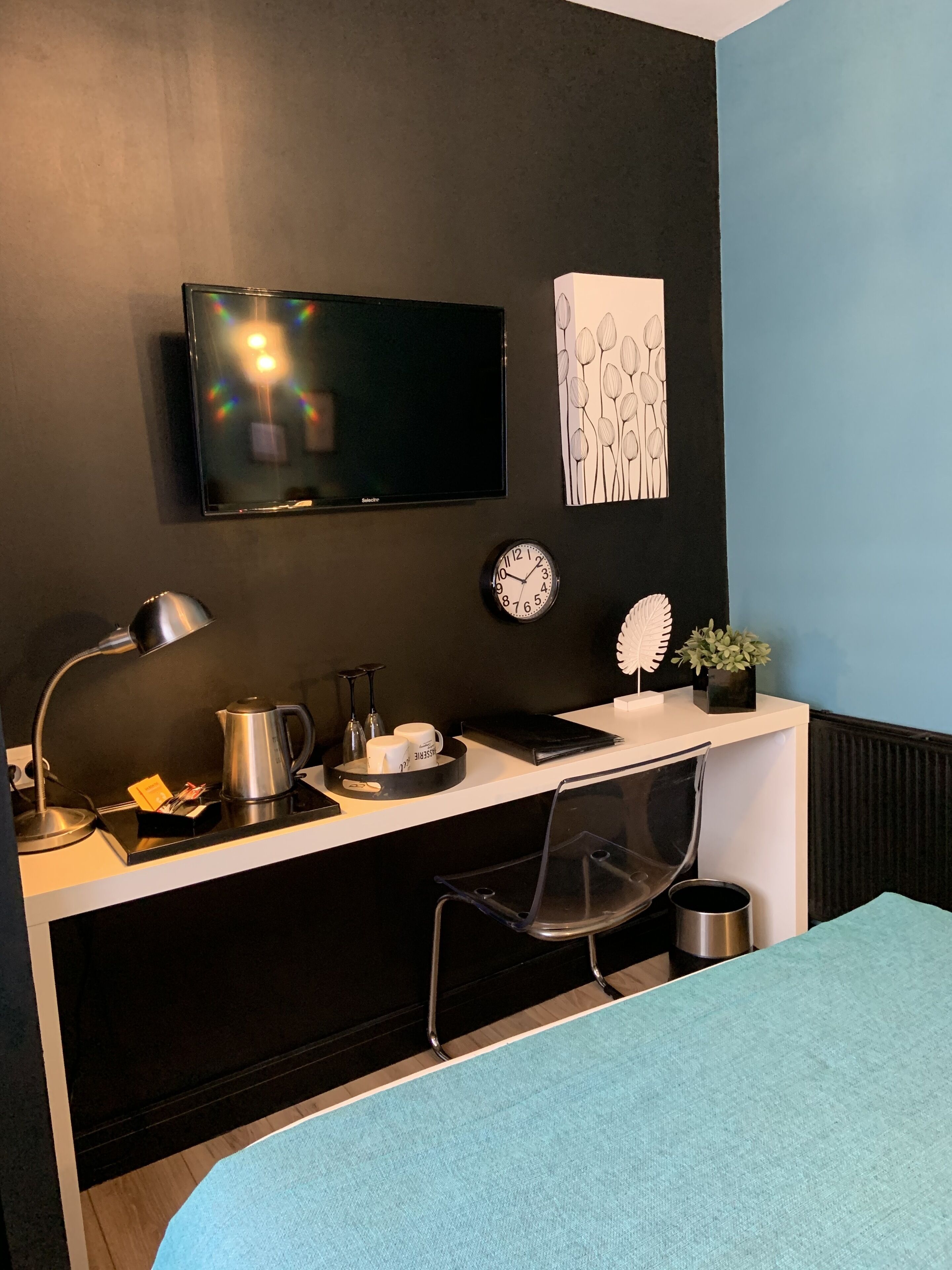 Moderni hotellihuone, jonka värimaailma on mustavalkoinen. Huoneessa on seinälle asennettu taulutelevisio, työpöytä, jossa on vedenkeitin, kuppeja ja kello, läpinäkyvä tuoli sekä pieni ruukkukasvi.