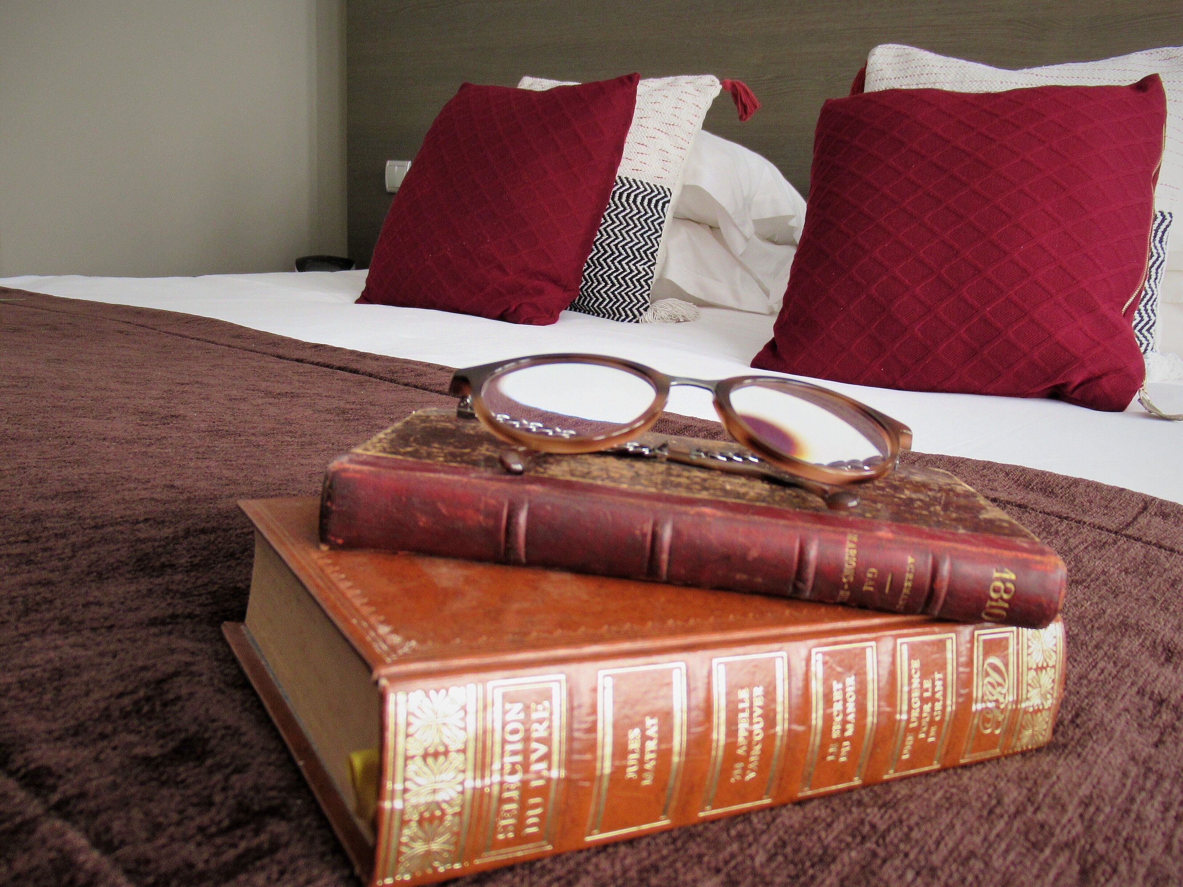 Una cama con una colcha blanca y dos almohadas rojas, un par de gafas y dos libros sobre una manta marrón.