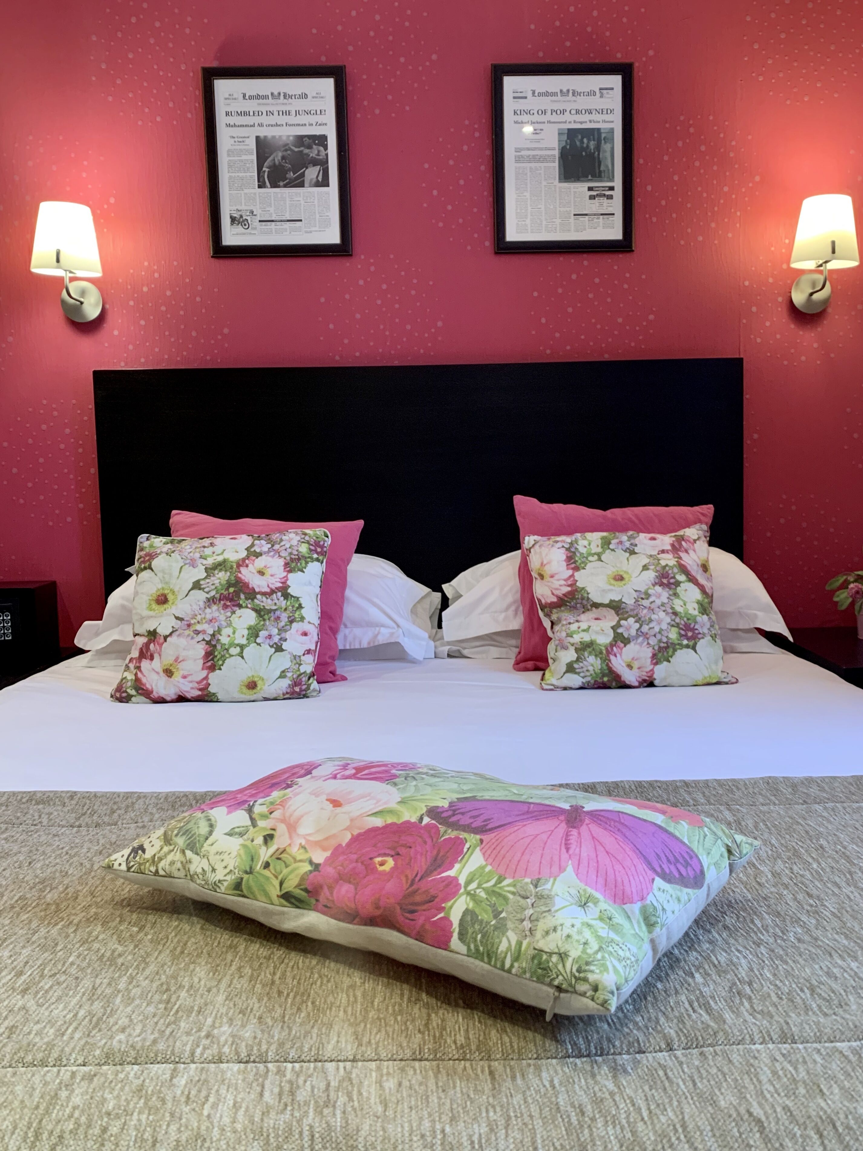 Ein Bett mit blumigen Kissen und passender Tagesdecke, ein schwarzes Kopfteil sowie zwei gerahmte Zeitungsartikel an der Wand.
