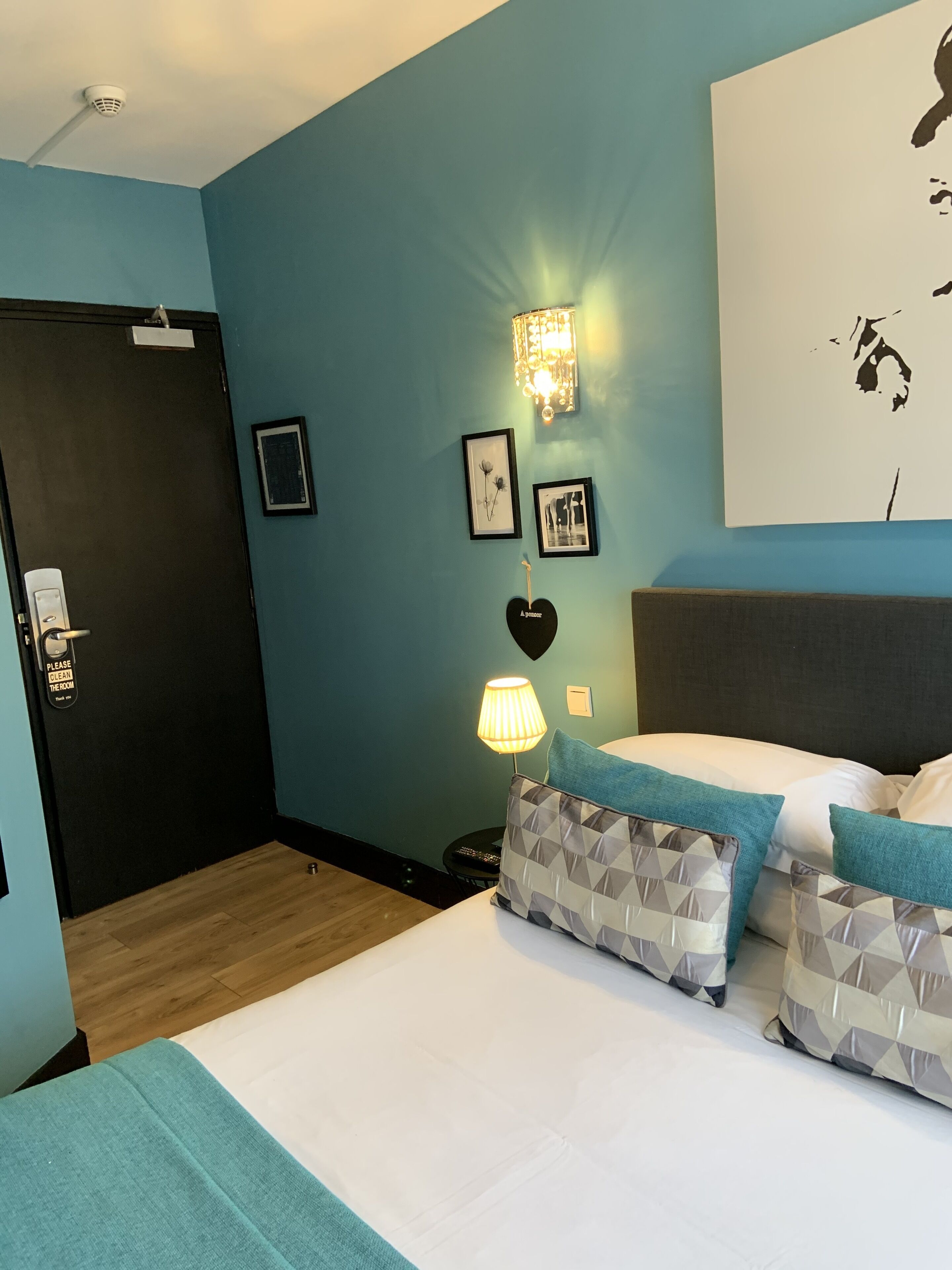 A hotel room with a bed, a nightstand with a lamp, a wall-mounted light fixture, and a door with a sign.