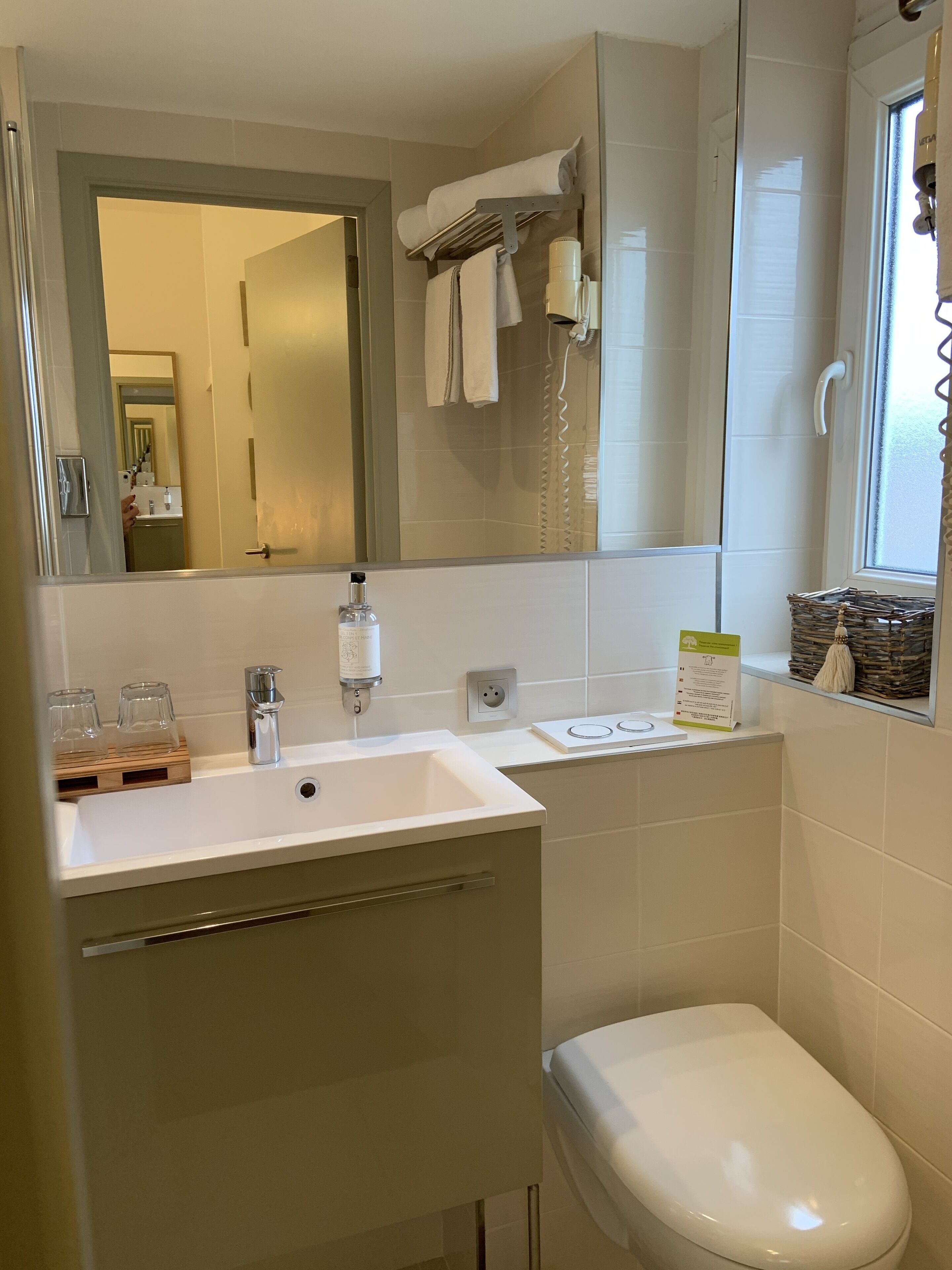 Een badkamer met een wit wastafel, spiegel en toilet. Boven het wastafel hangt een handdoekrek en er is een raam met uitzicht op buiten.