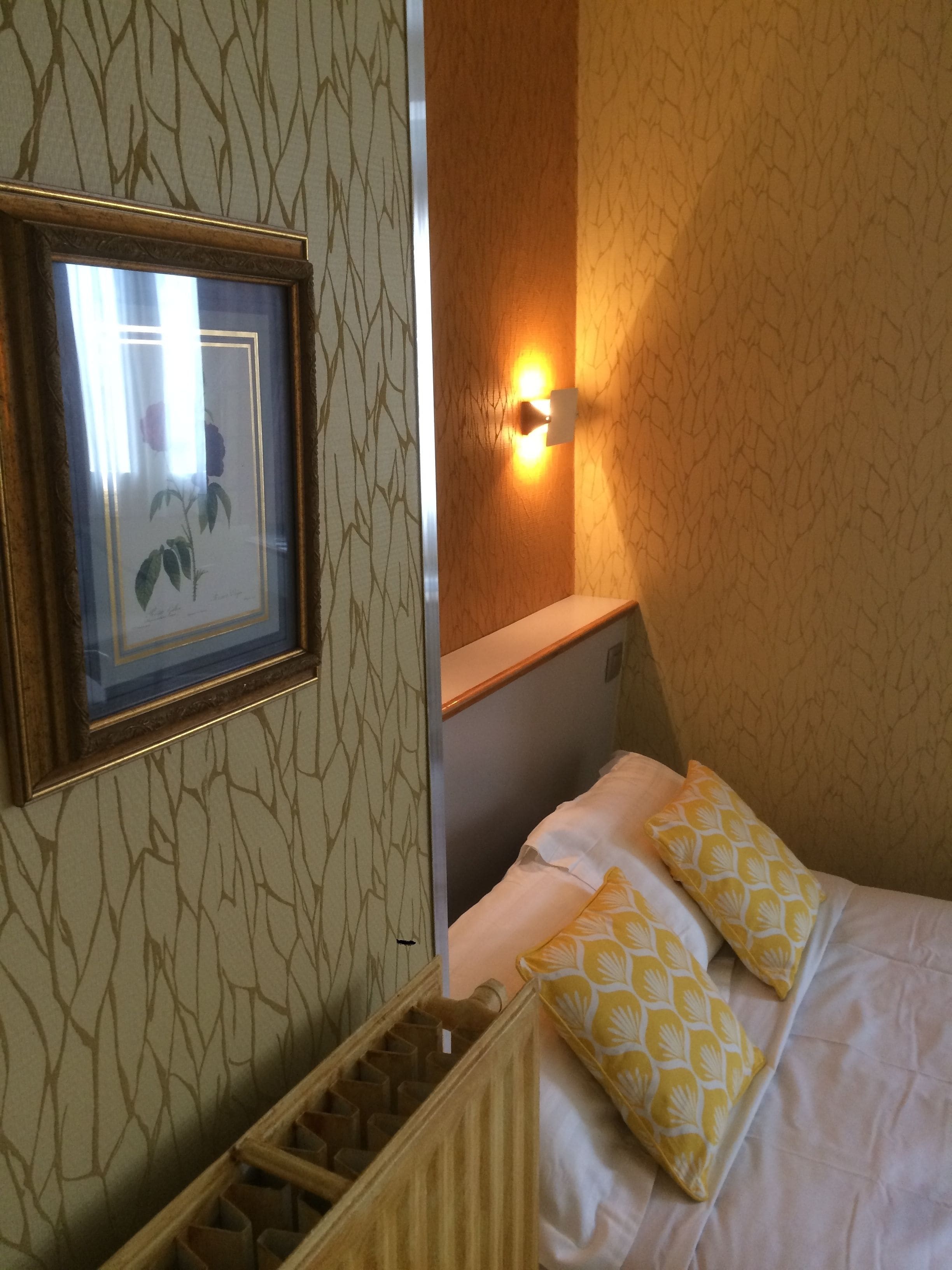 A hotel room with a framed picture on the wall, a bedside lamp, a bed with white bedding and yellow pillows, and a radiator.