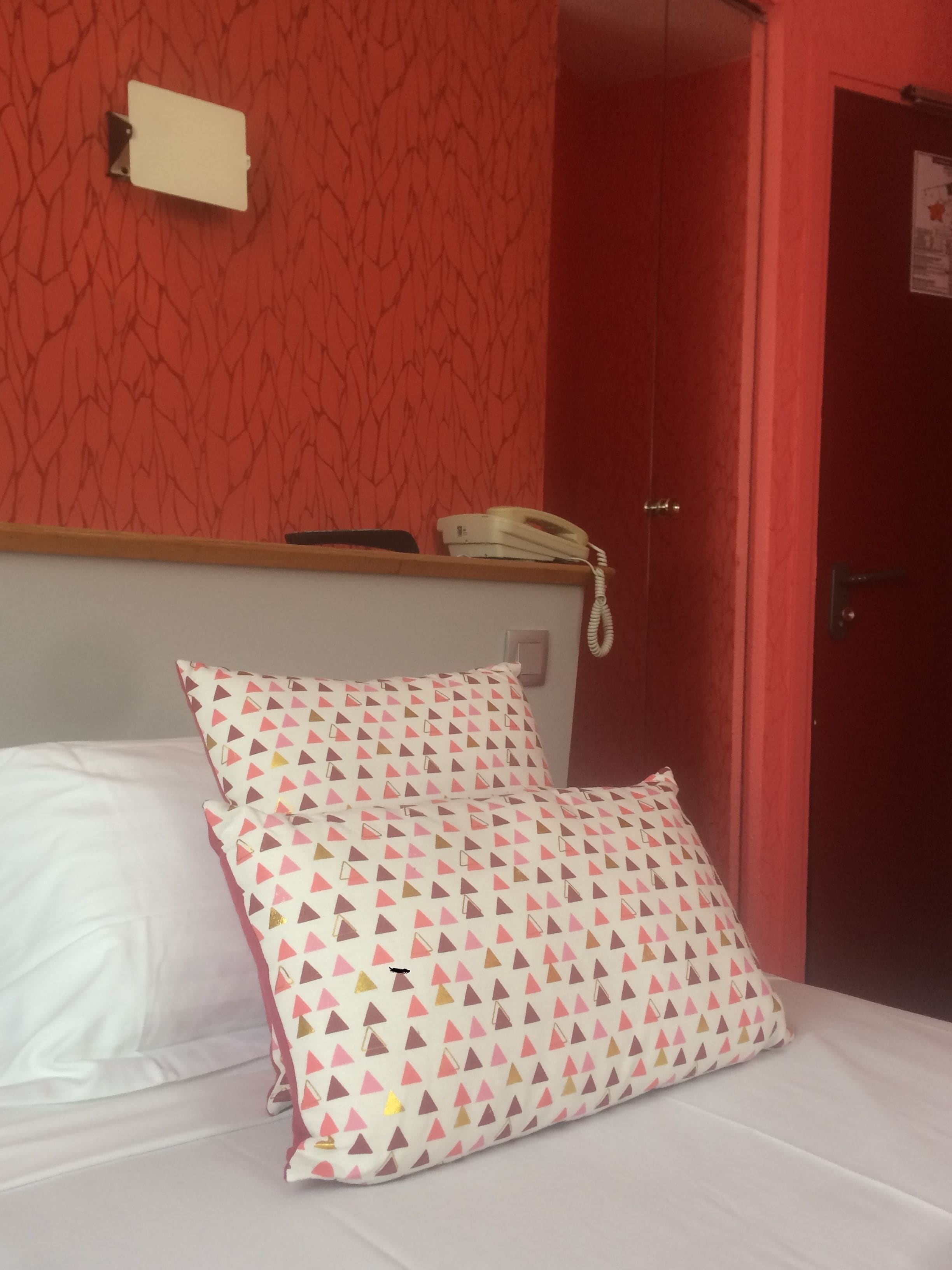 A bed with white and pink patterned pillows, a bedside telephone, and a red patterned wall.