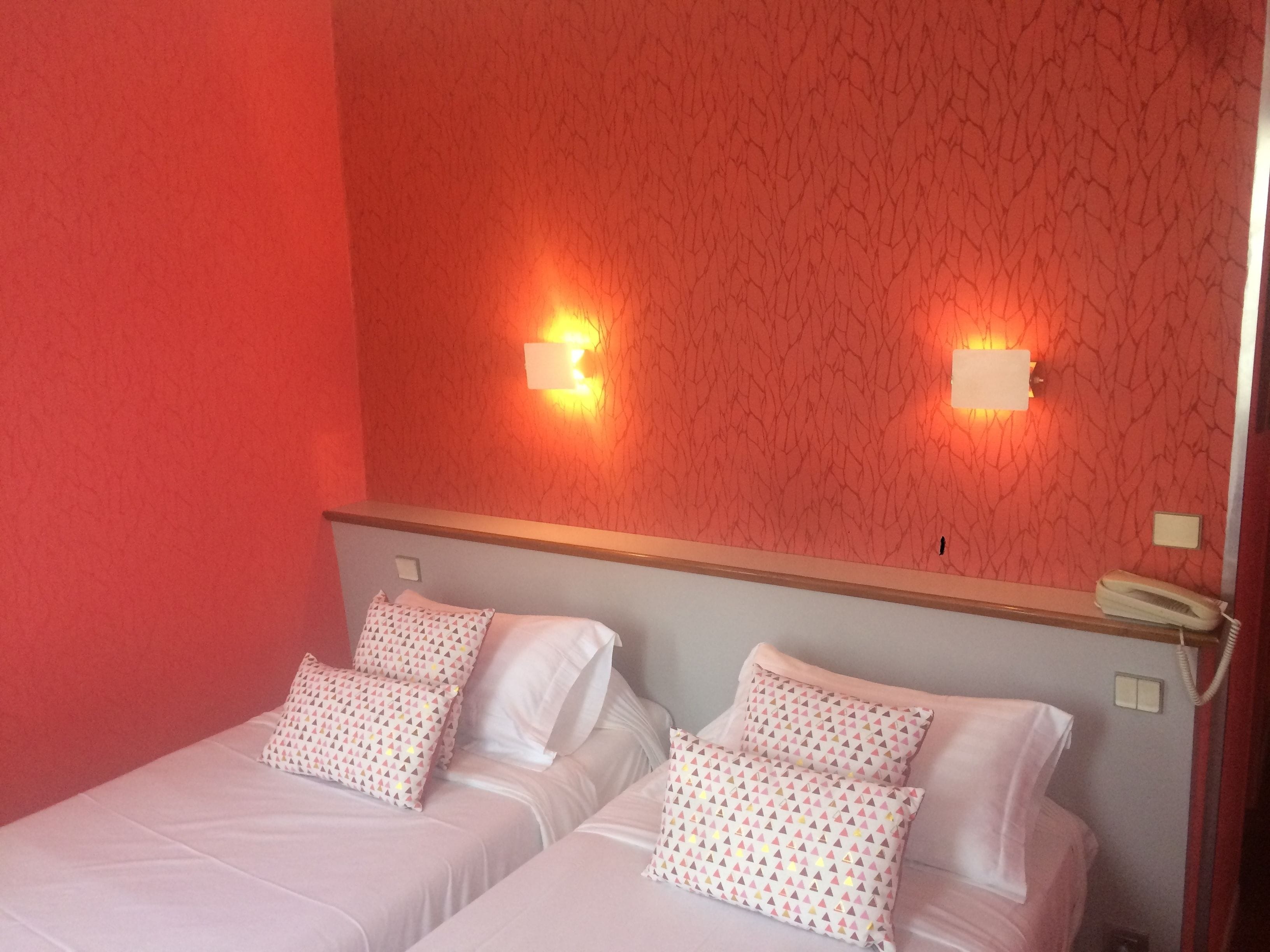 A hotel room with two beds, each with white bedding and patterned pillows. There are wall-mounted light fixtures on the red wall.