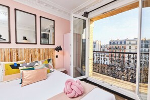 Une chambre à coucher avec un lit, des oreillers, un miroir et une fenêtre donnant sur des immeubles.